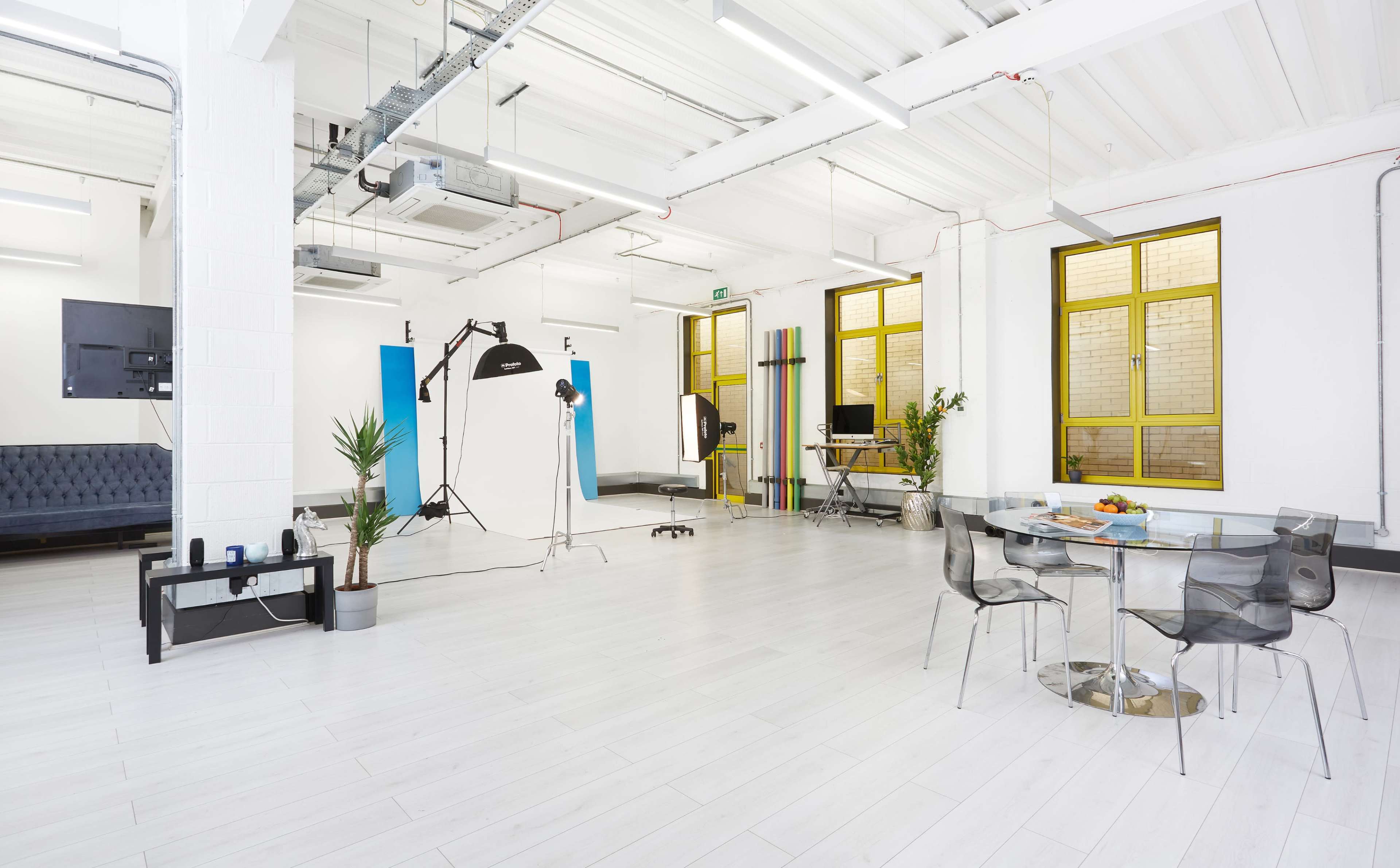 A spacious, well-lit studio features white walls, a gray sofa, and a glass dining table surrounded by transparent chairs, with a photography setup in one corner.