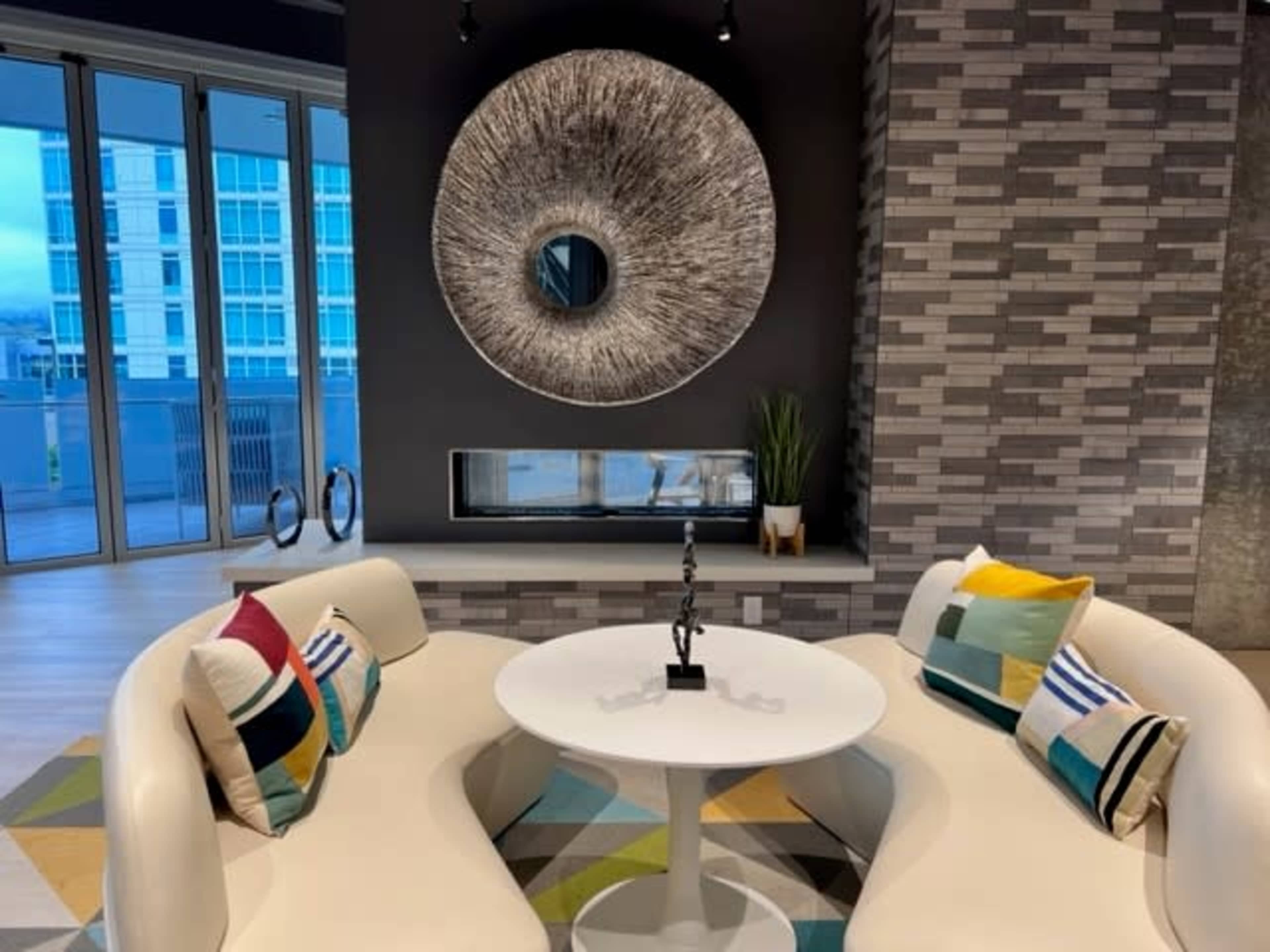 A modern lounge area with two curved sofas facing a round table, a decorative wall mirror, and a fireplace beneath a gray tiled wall.