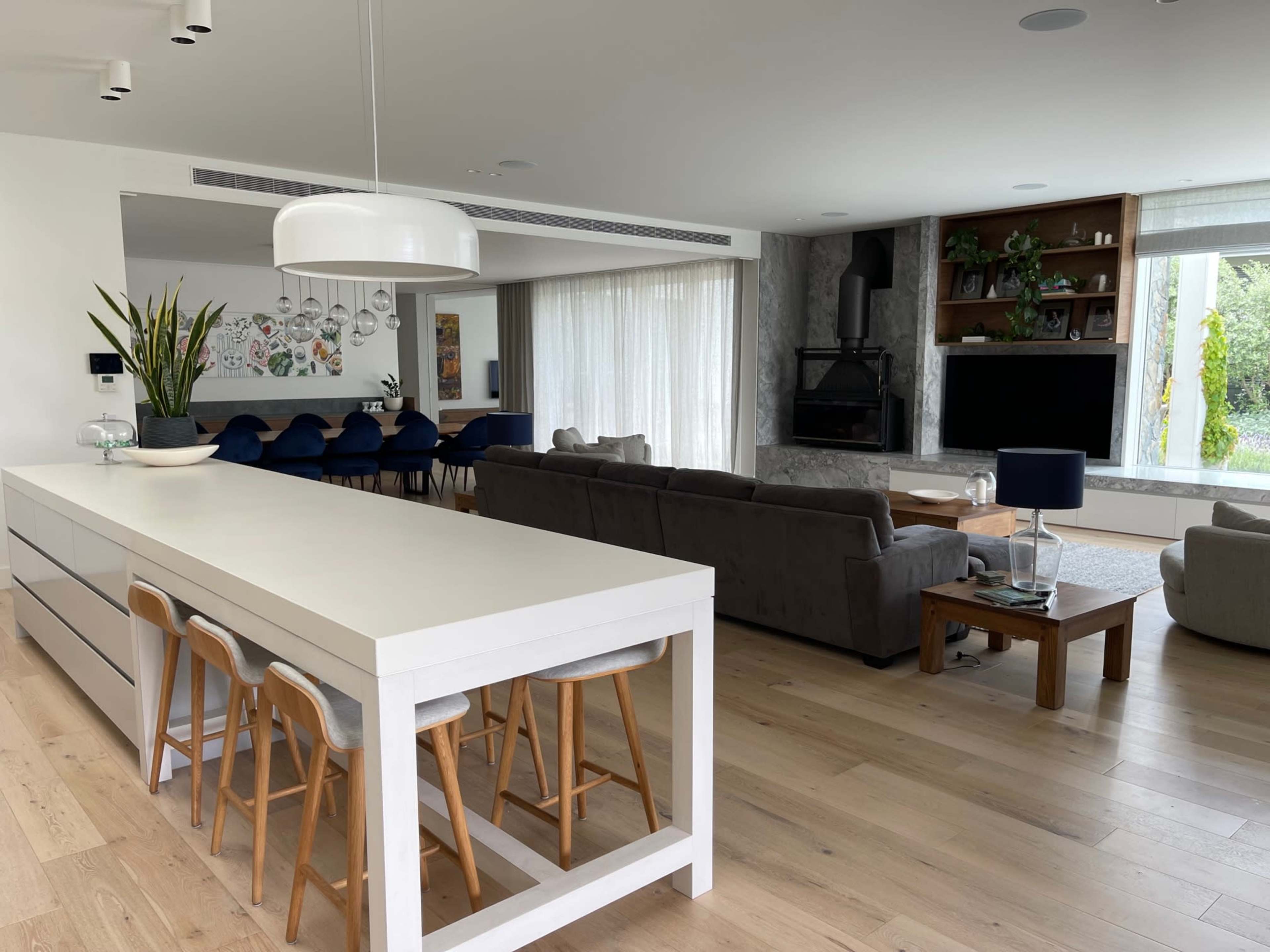 A modern open-concept living space features a kitchen island with bar stools, a dining area, and a living room with large windows and a fireplace.
