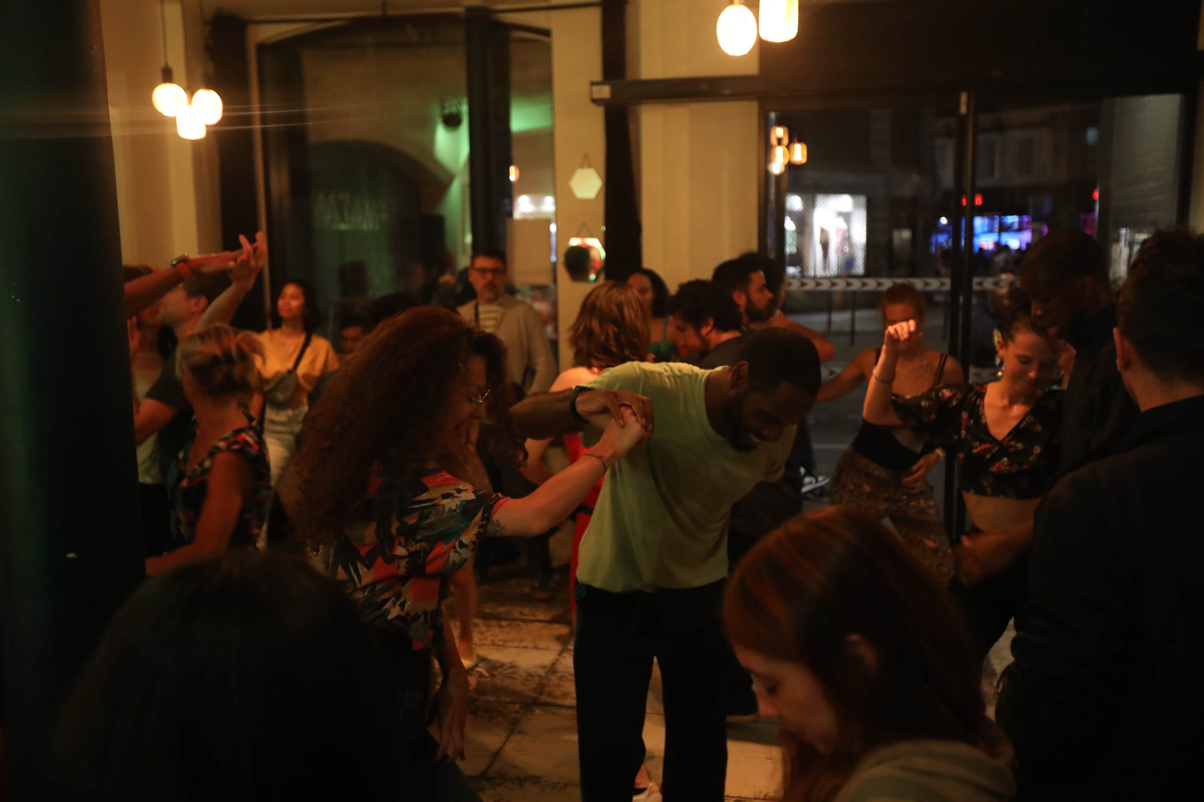 A diverse group of people dances energetically together in a warmly lit venue with large windows.