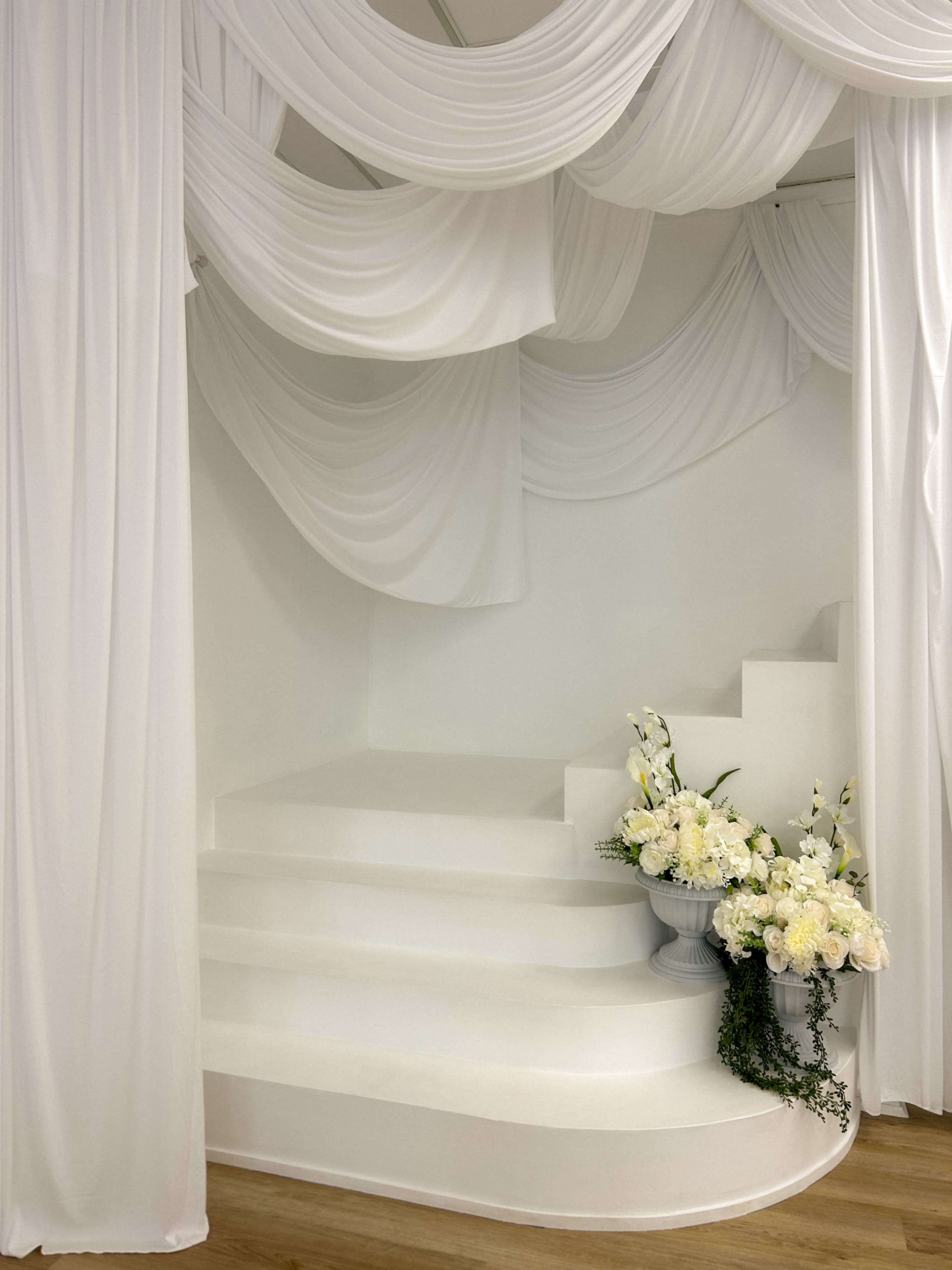 The image features a white, draped fabric backdrop over a set of white stairs, accompanied by floral arrangements in a vase on the side.
