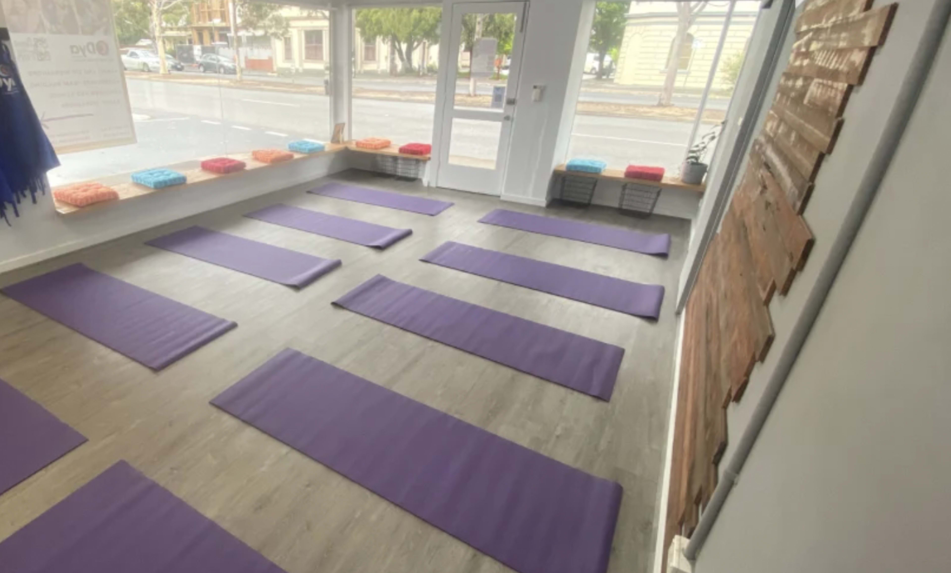 The image shows a yoga studio with several purple mats arranged in rows on a wooden floor, and colorful cushions lined up on a window ledge.