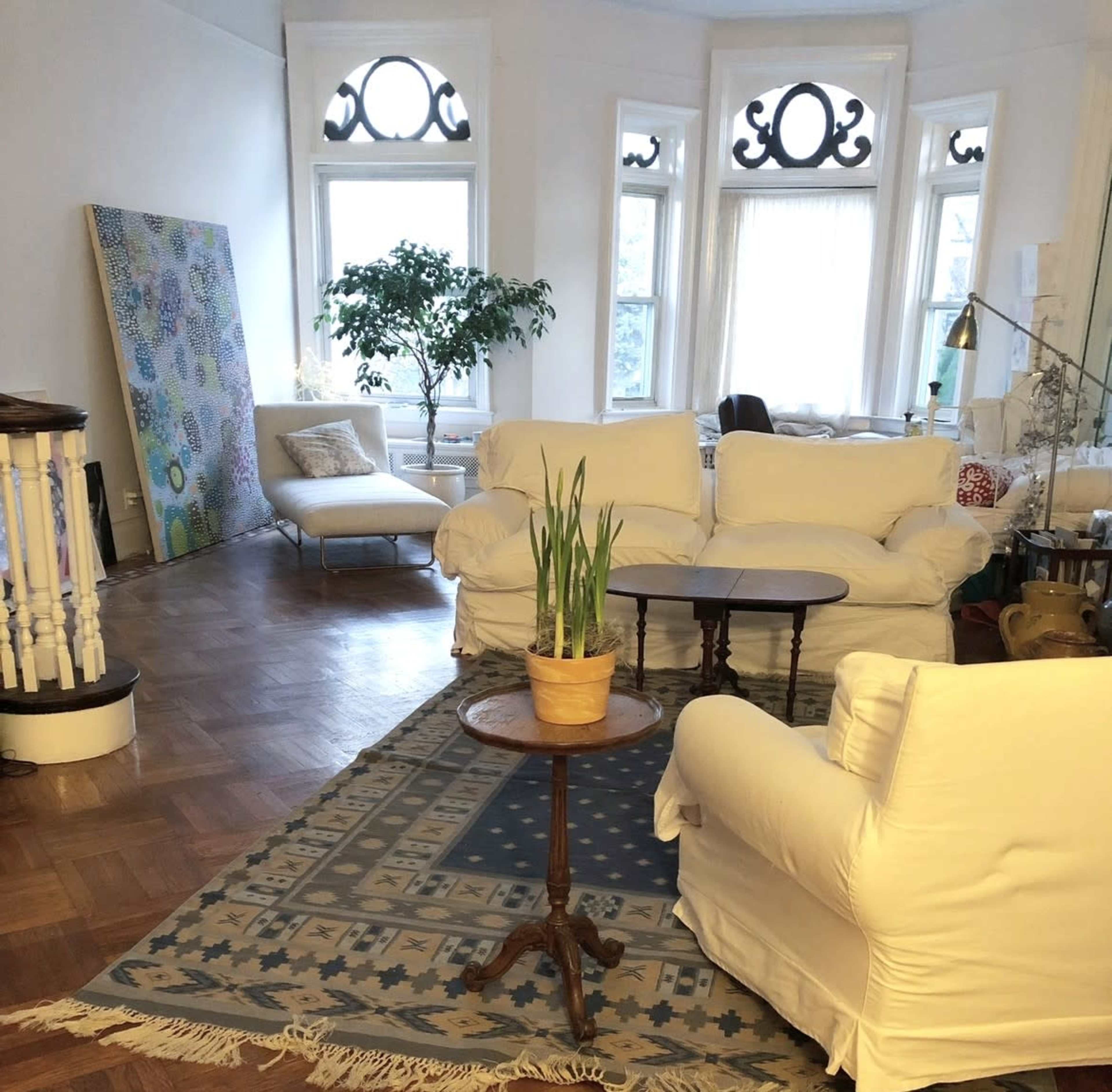 The image shows a well-furnished living room with white upholstered chairs, a small wooden table, and a potted plant, along with large windows that allow natural light to fill the space.