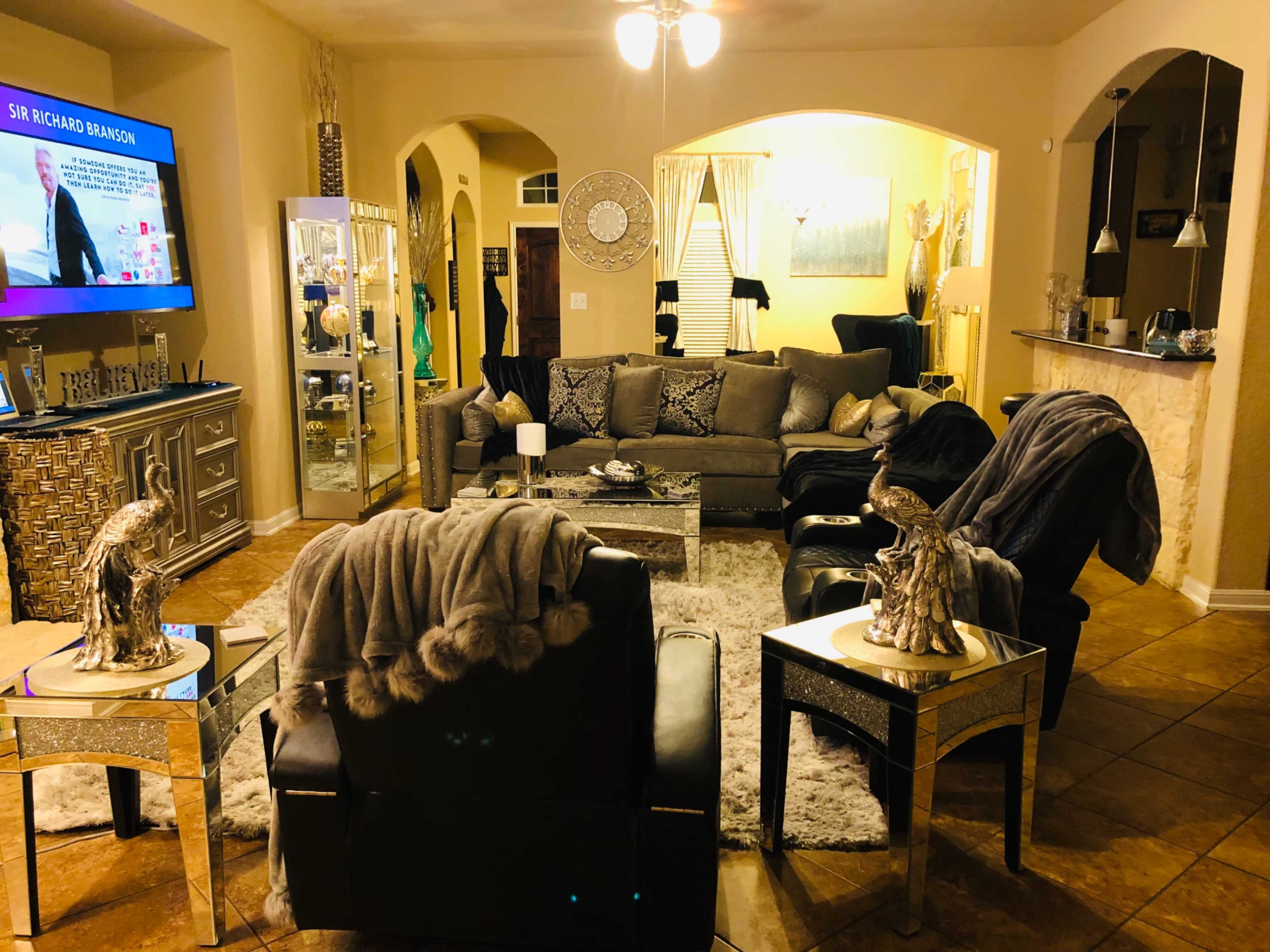 The living room features a large gray sectional sofa, two black chairs, a glass coffee table, and a television mounted on the wall.
