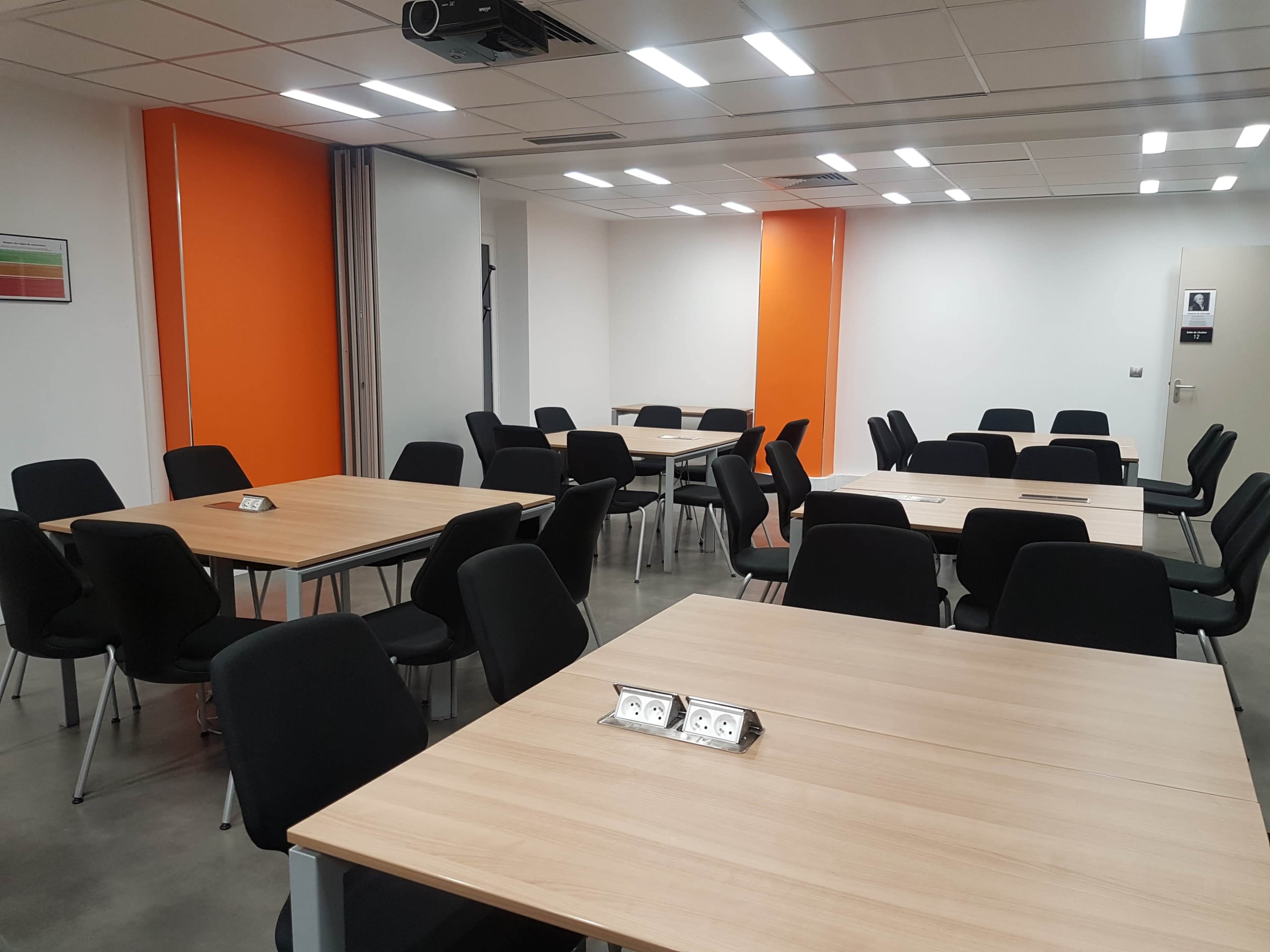 A modern conference room features several rectangular tables with black chairs arranged around them, against a backdrop of white walls with an orange accent wall.