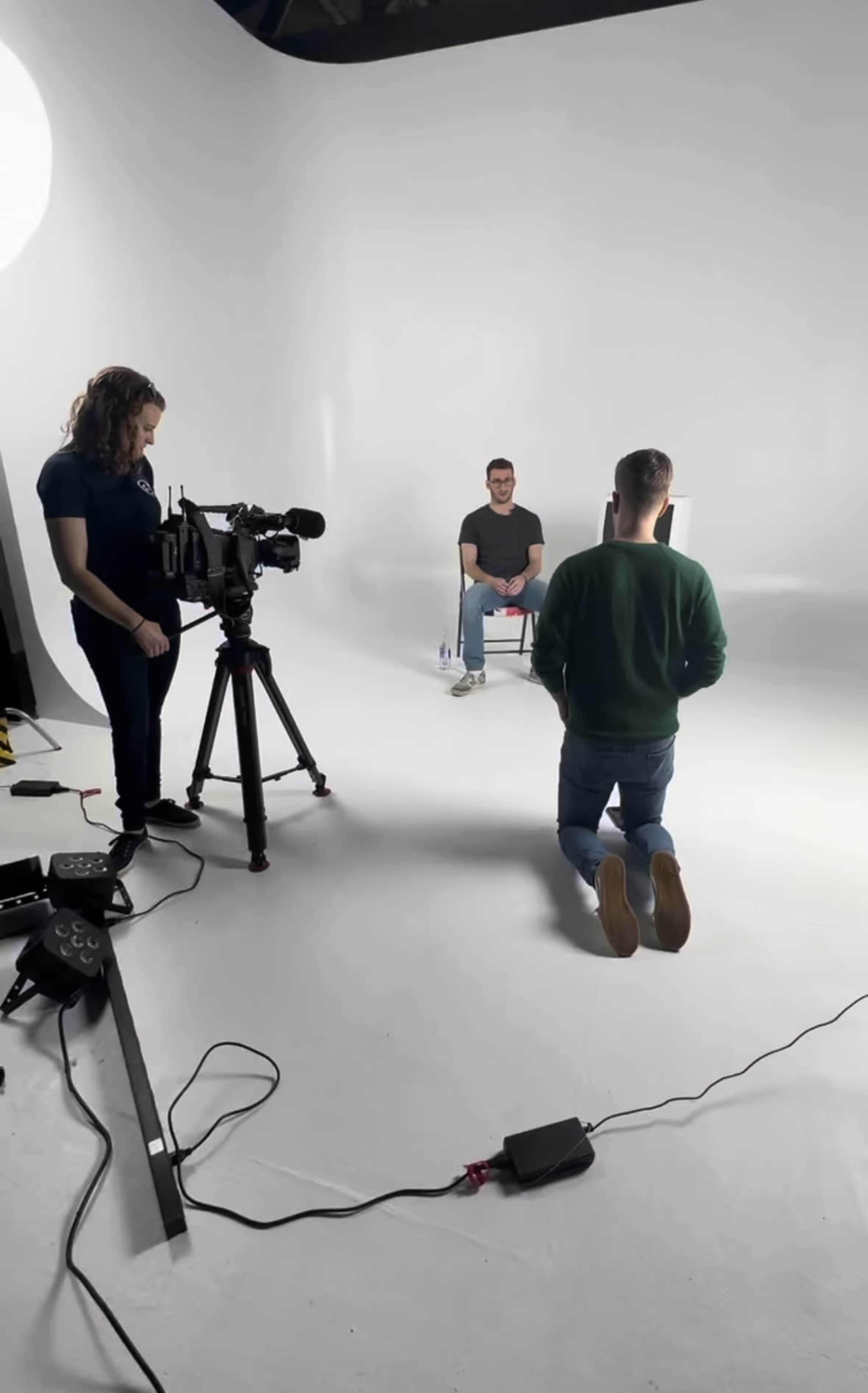 A film crew sets up in a studio with a seated interviewee in a chair, while a person kneels on the floor facing the camera.