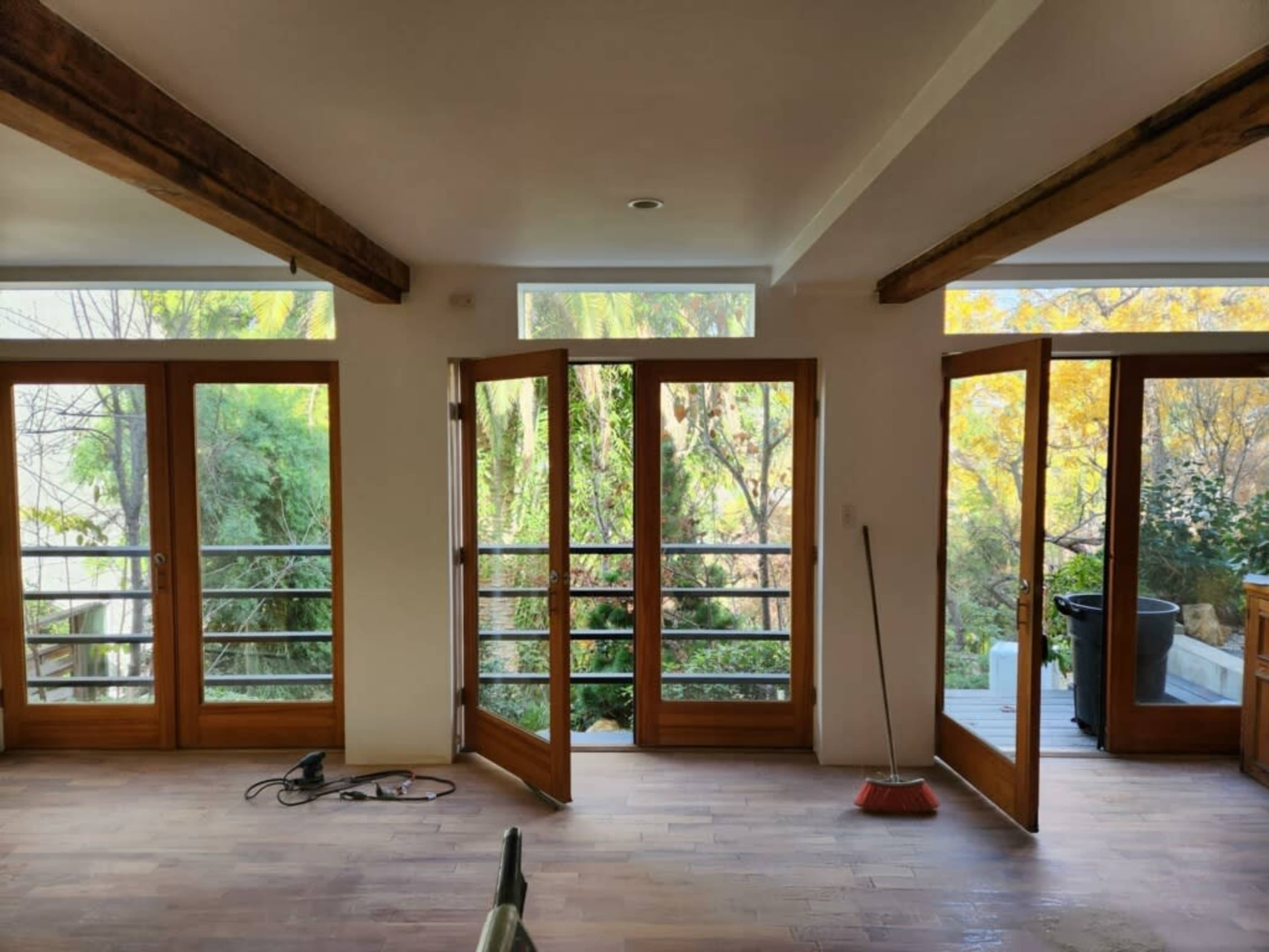 The room features large wooden doors that open to a view of greenery outside, with a broom and electrical cord on the floor.