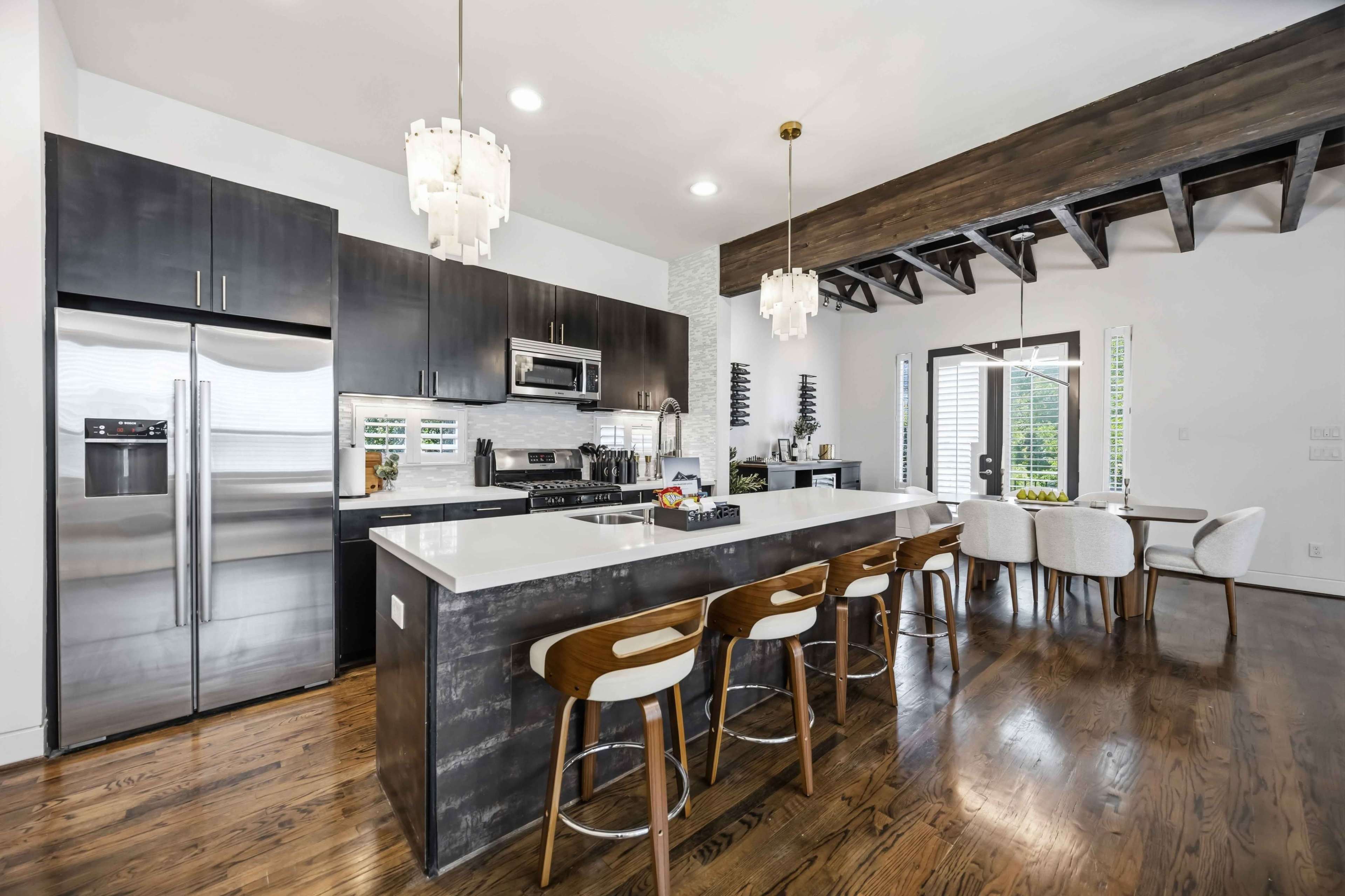 The image shows a modern kitchen with dark cabinetry, a large island with seating, stainless steel appliances, and a dining area featuring a circular table and chairs.