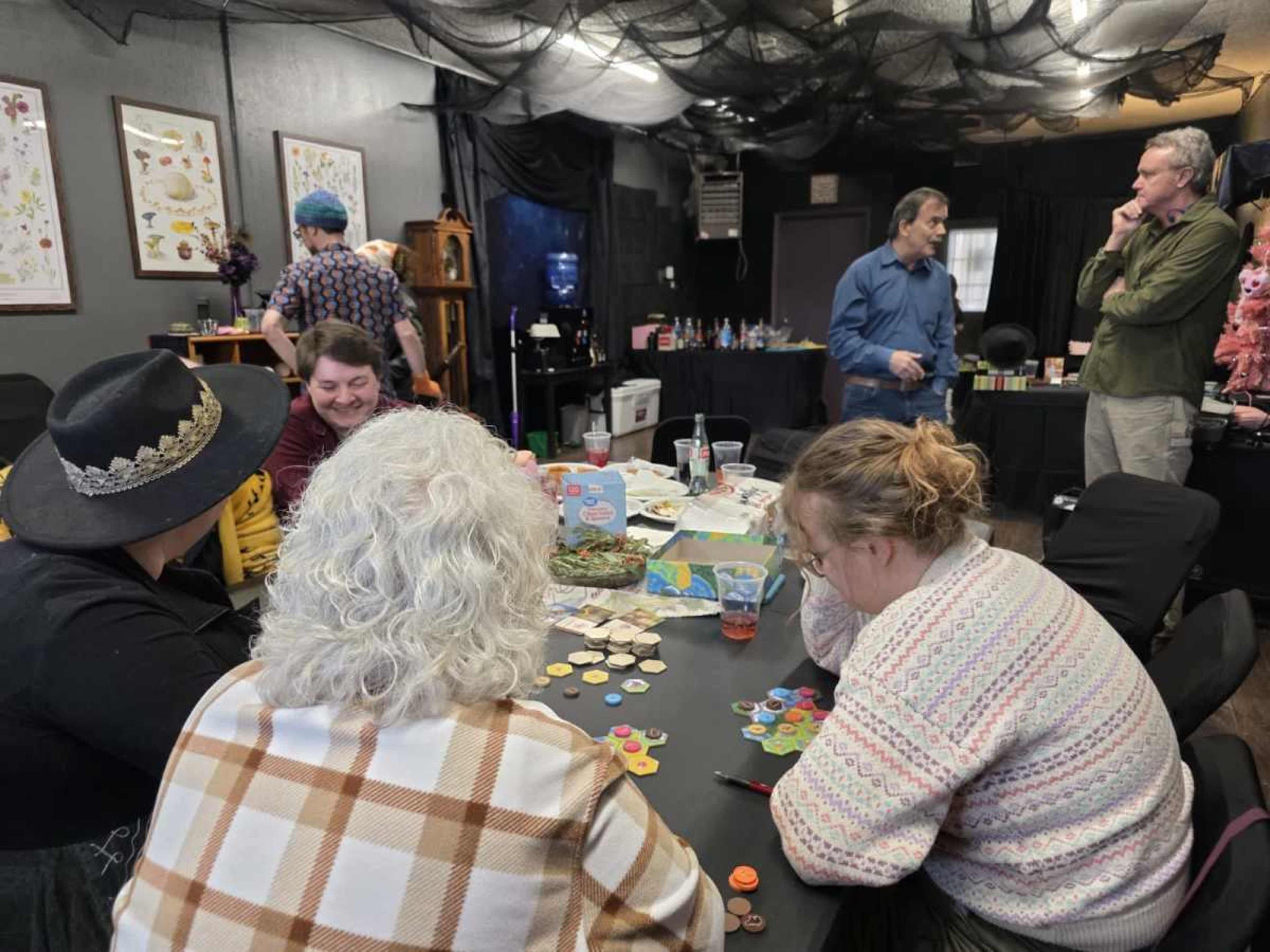 A group of people is playing board games at a table, while others are socializing in a decorated room with dim lighting.