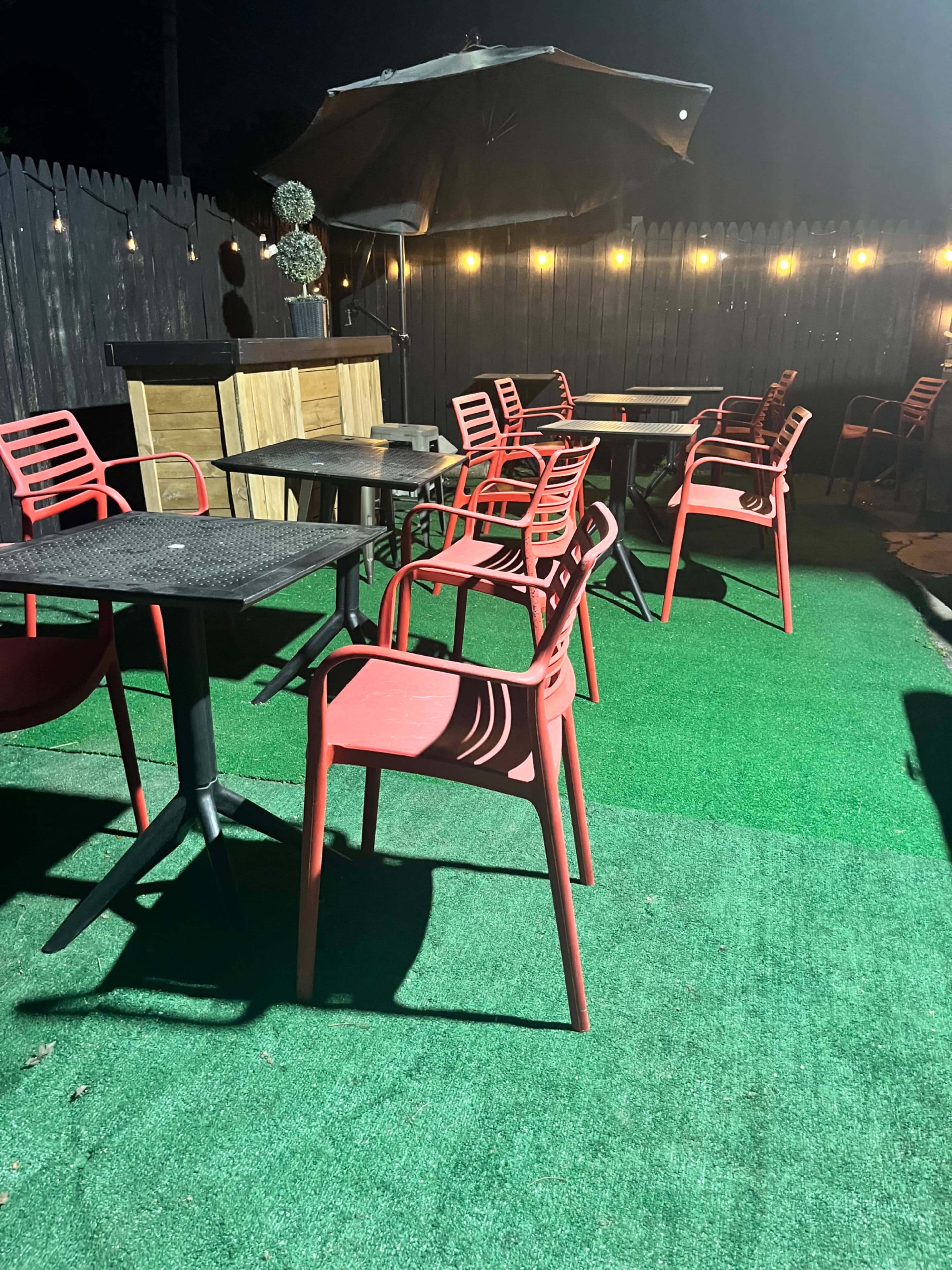 The image shows an outdoor seating area at night, featuring several red chairs around black tables on a green turf surface, with dim lighting from overhead fixtures.