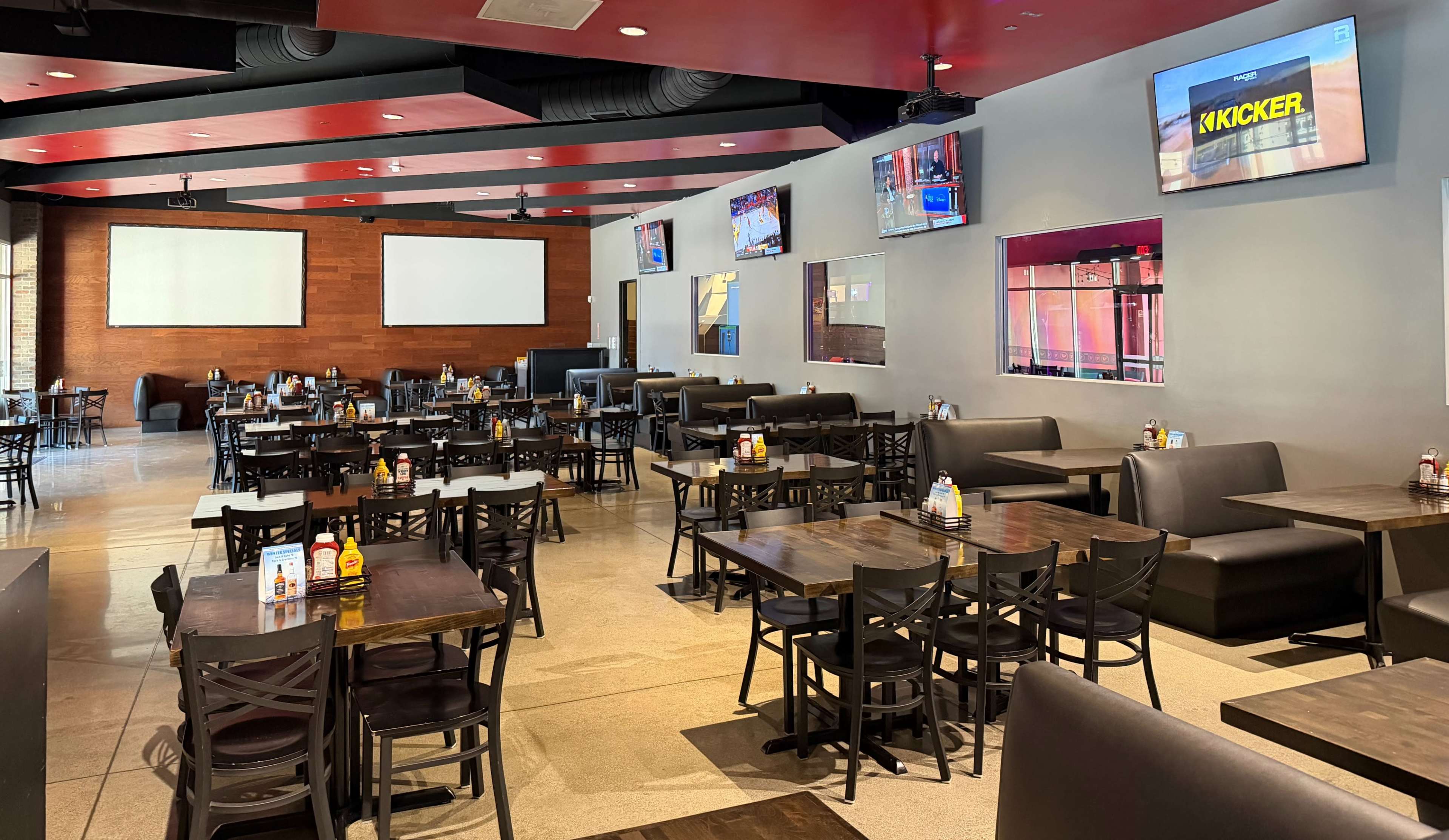 The image shows an interior of a restaurant with numerous tables and chairs arranged in a spacious layout, featuring multiple televisions mounted on the walls.