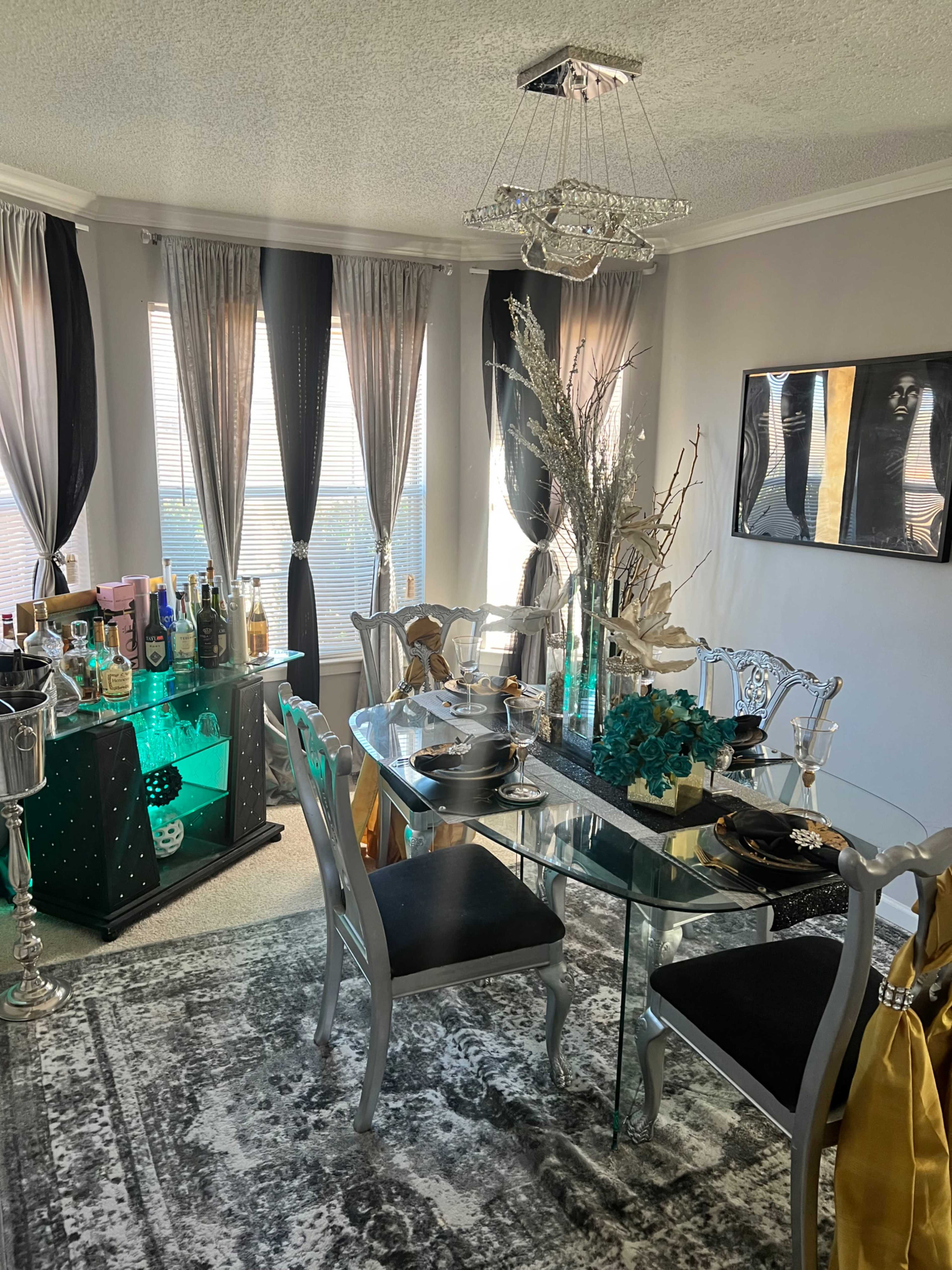 A modern dining room featuring a glass dining table with black and silver chairs, a bar cart stocked with various bottles, and elegant decor elements, including a chandelier and table settings.