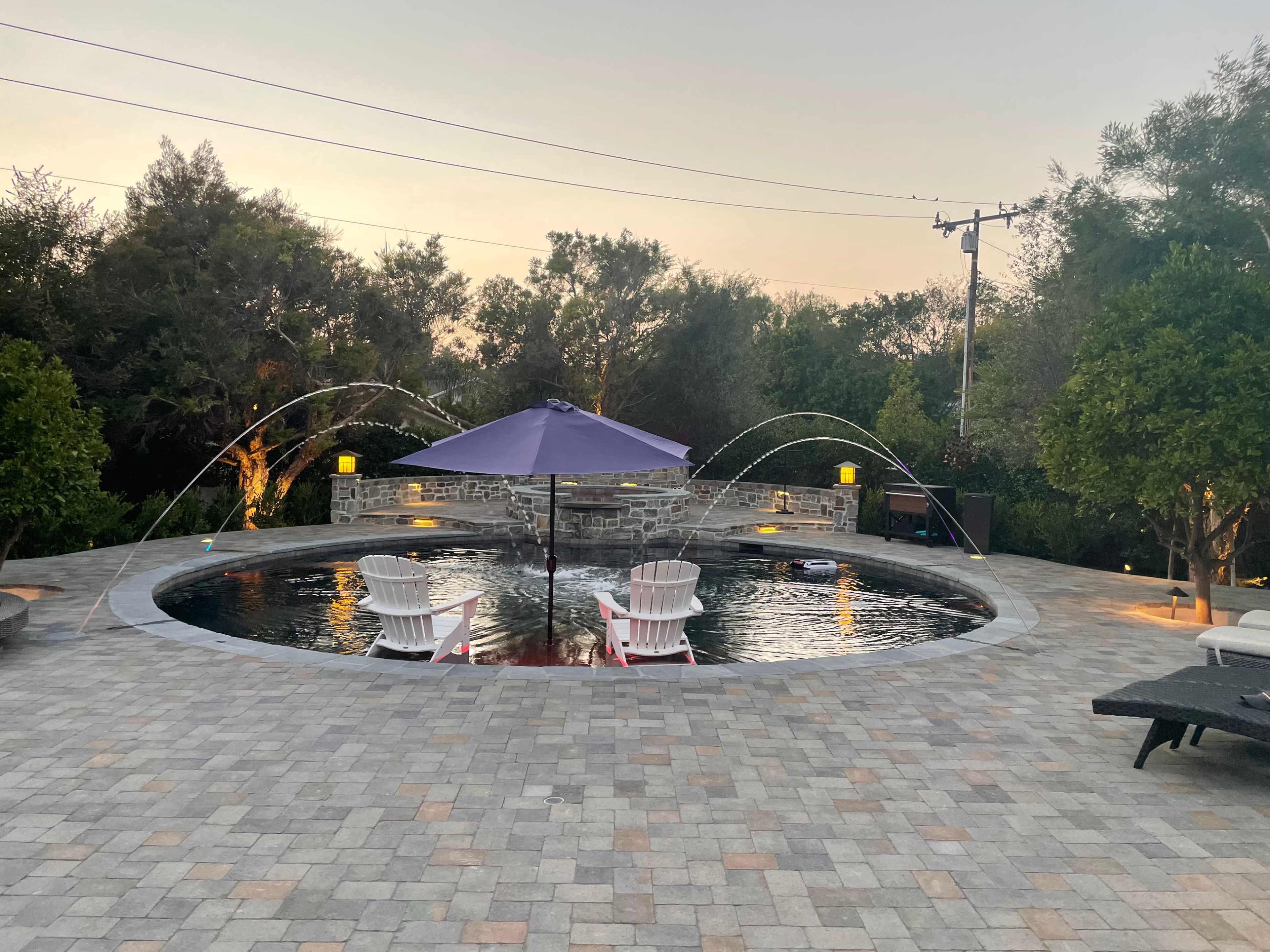 The image shows a circular swimming pool surrounded by stone pavers, with white lounge chairs and a purple umbrella, and two water fountains spraying into the pool at dusk.