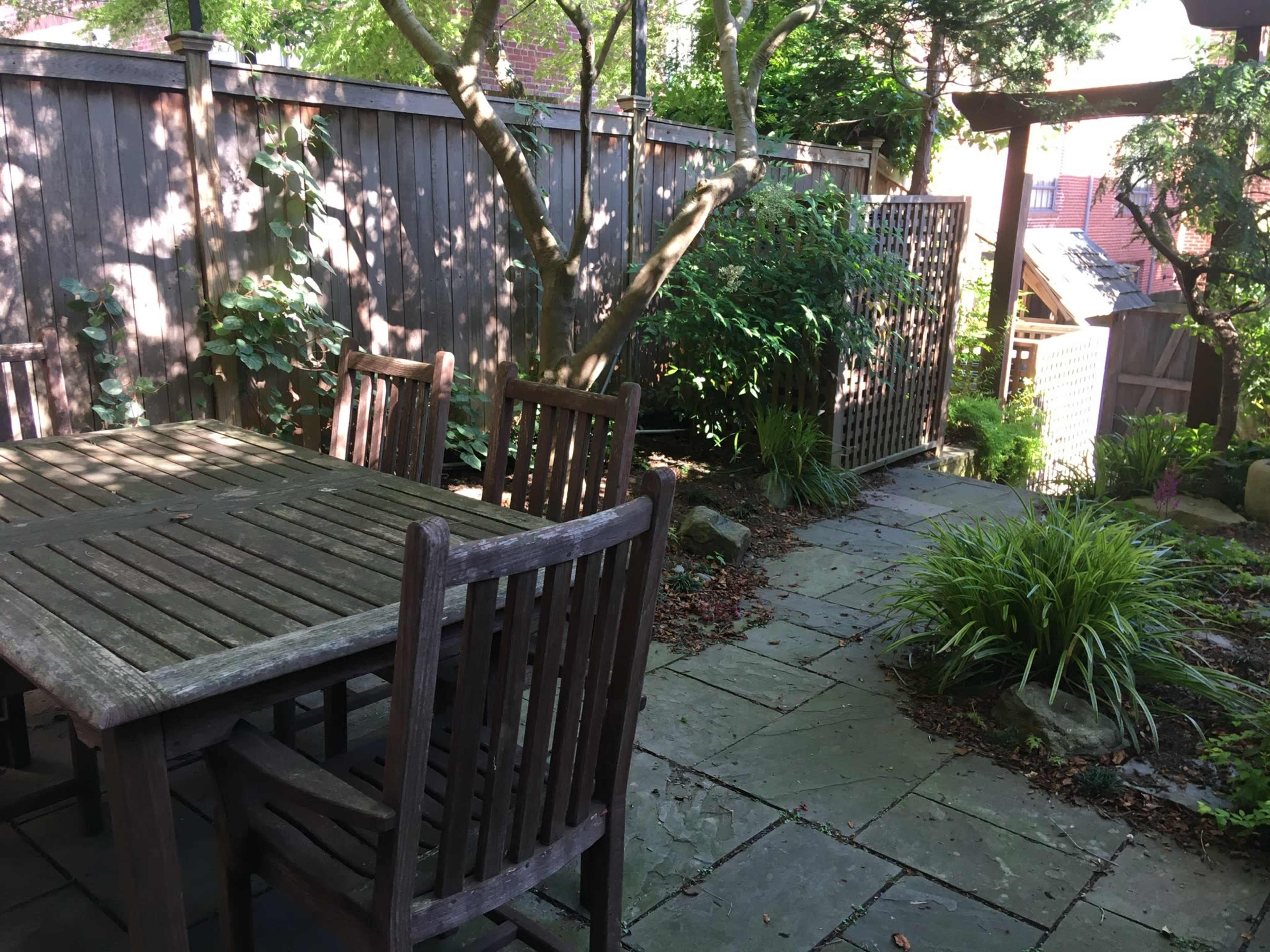 A wooden table with chairs is set on a stone patio surrounded by greenery and a fenced area.