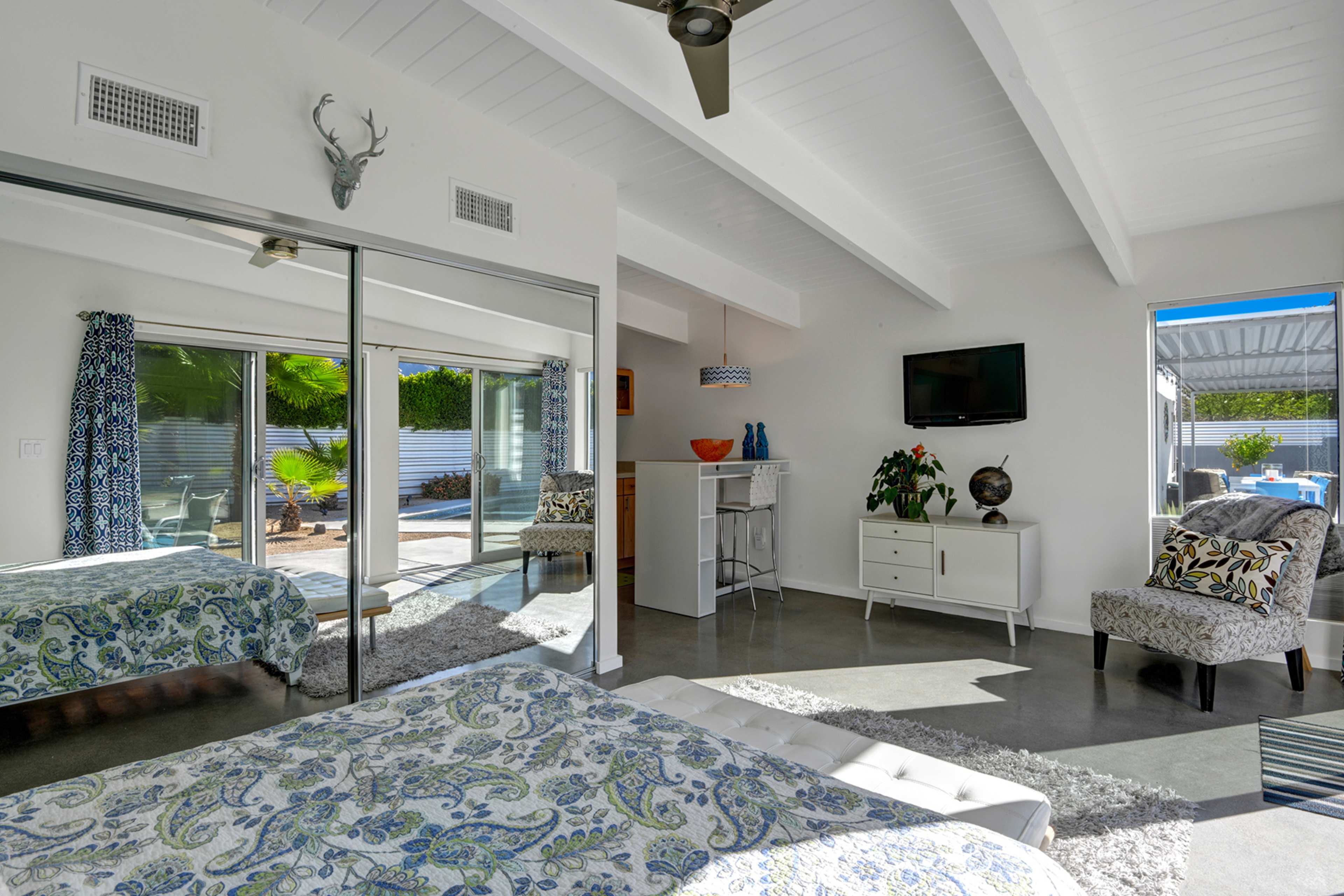 The image shows a bright bedroom with a large mirror, modern furniture, and sliding glass doors leading to an outdoor space.
