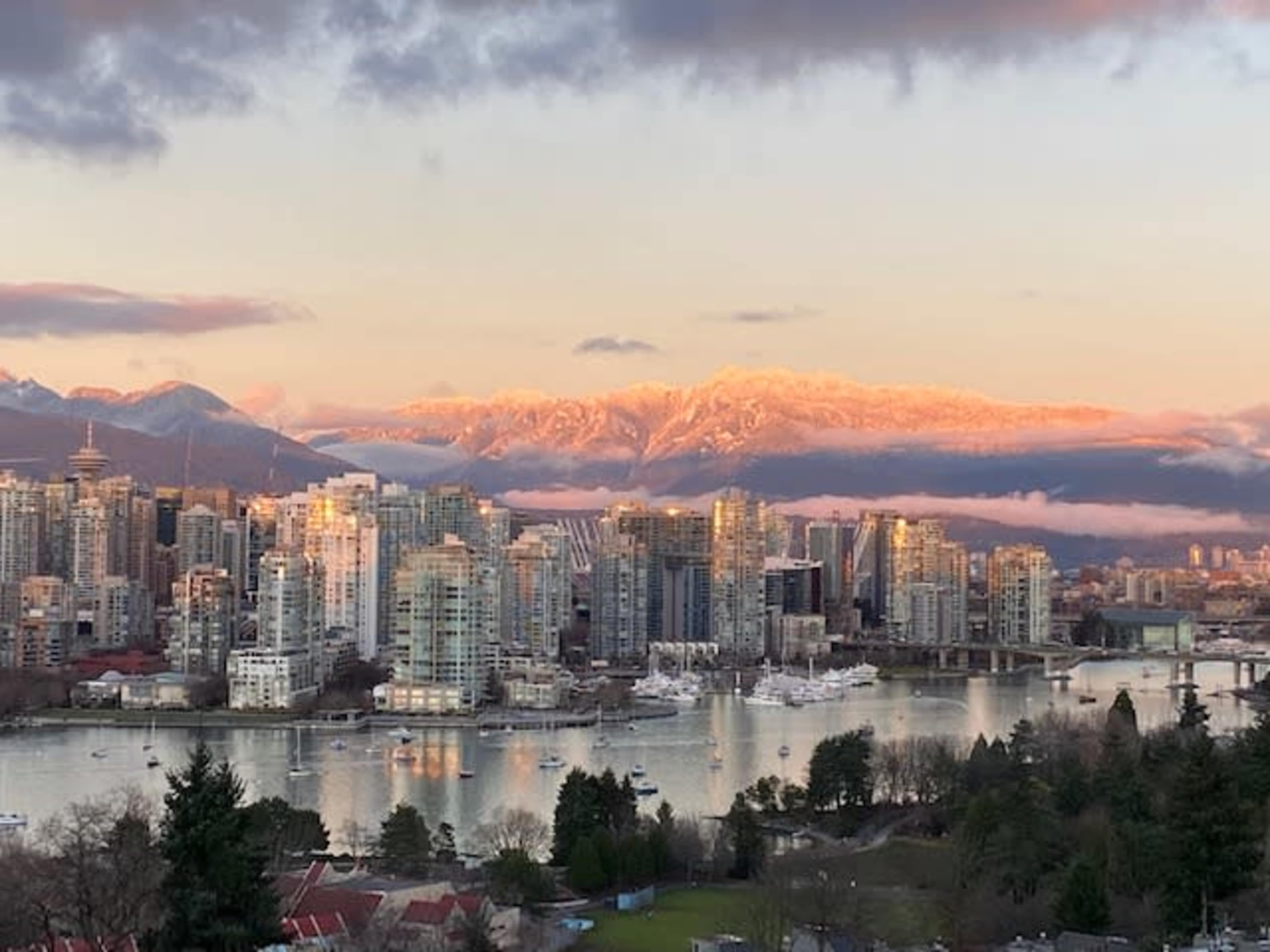 A city skyline with tall buildings along a river, framed by snow-capped mountains under a colorful sky at sunset.
