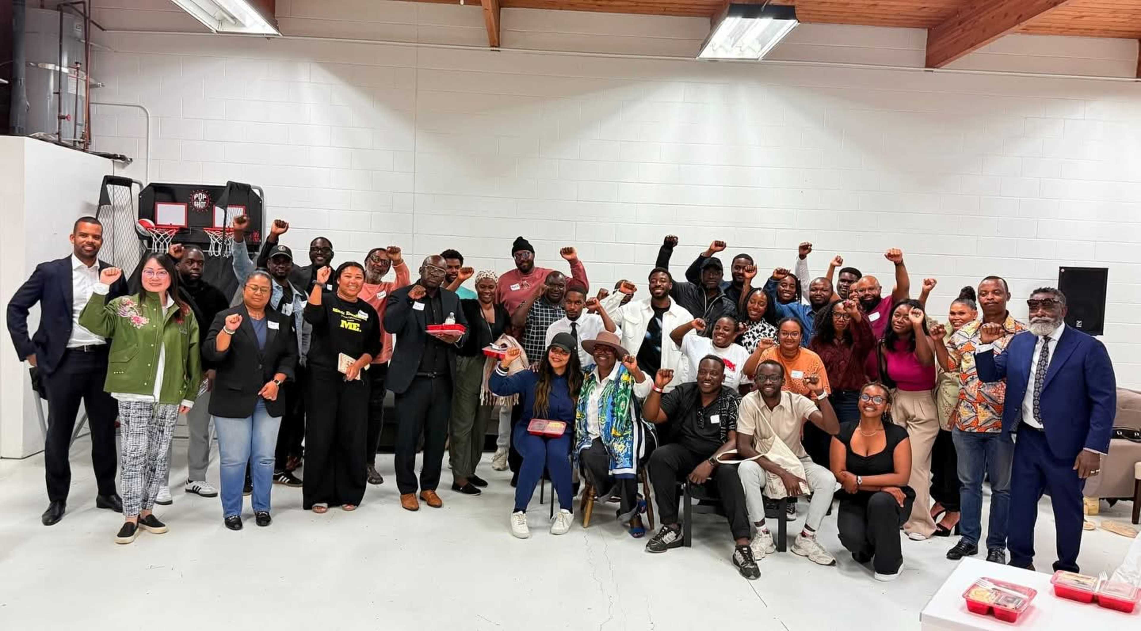 A large group of diverse individuals is gathered in a spacious indoor setting, with many smiling and raising their fists in a gesture of solidarity.