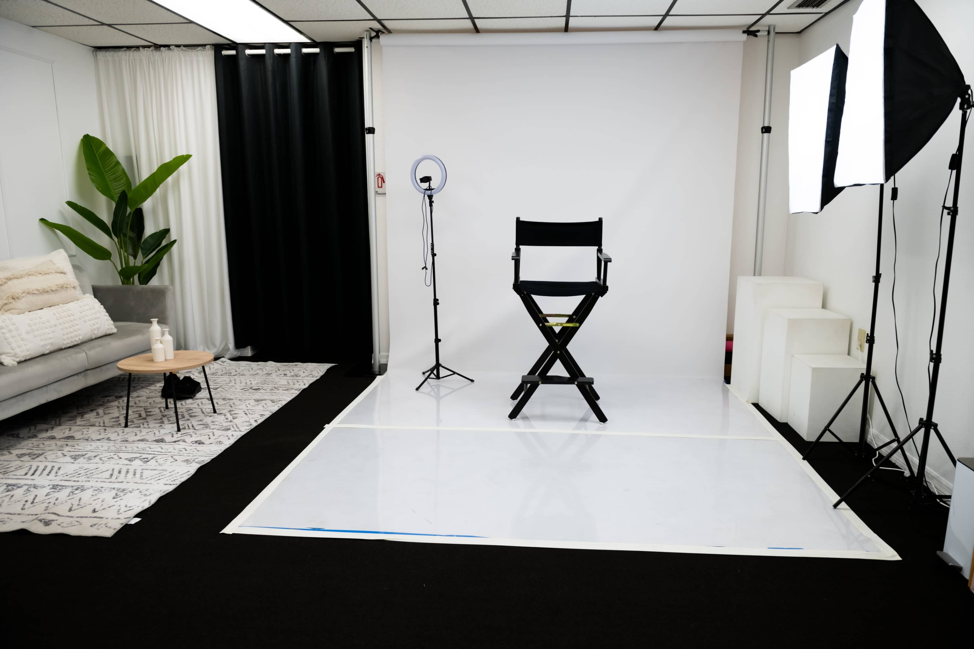 The image shows a minimalist studio setup with a director's chair in front of a plain backdrop, surrounded by lighting equipment and a plant.