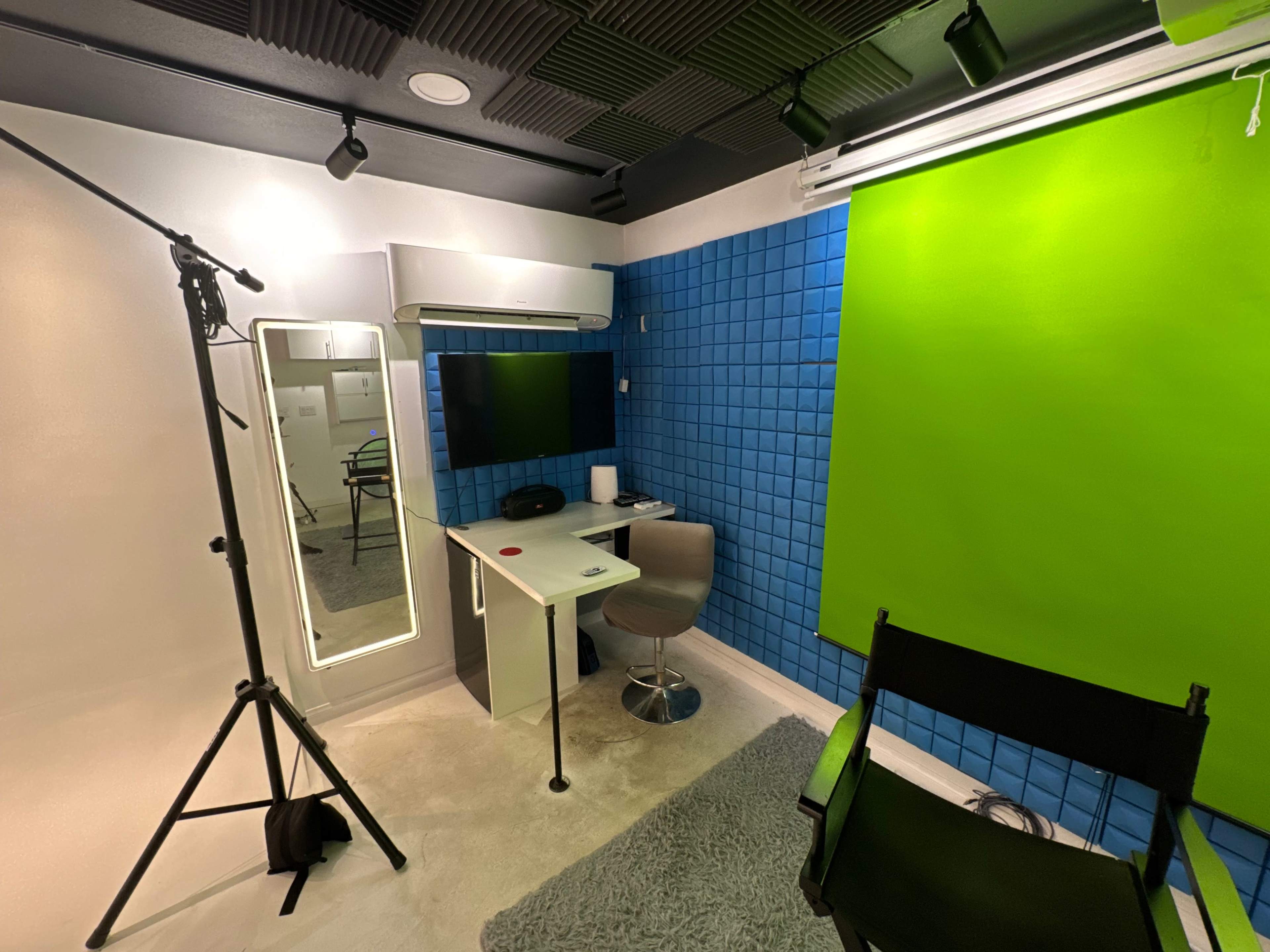 The image shows a recording setup featuring a green screen, a desk with a mirror, a TV mounted on the wall, and a director's chair in a brightly lit room.