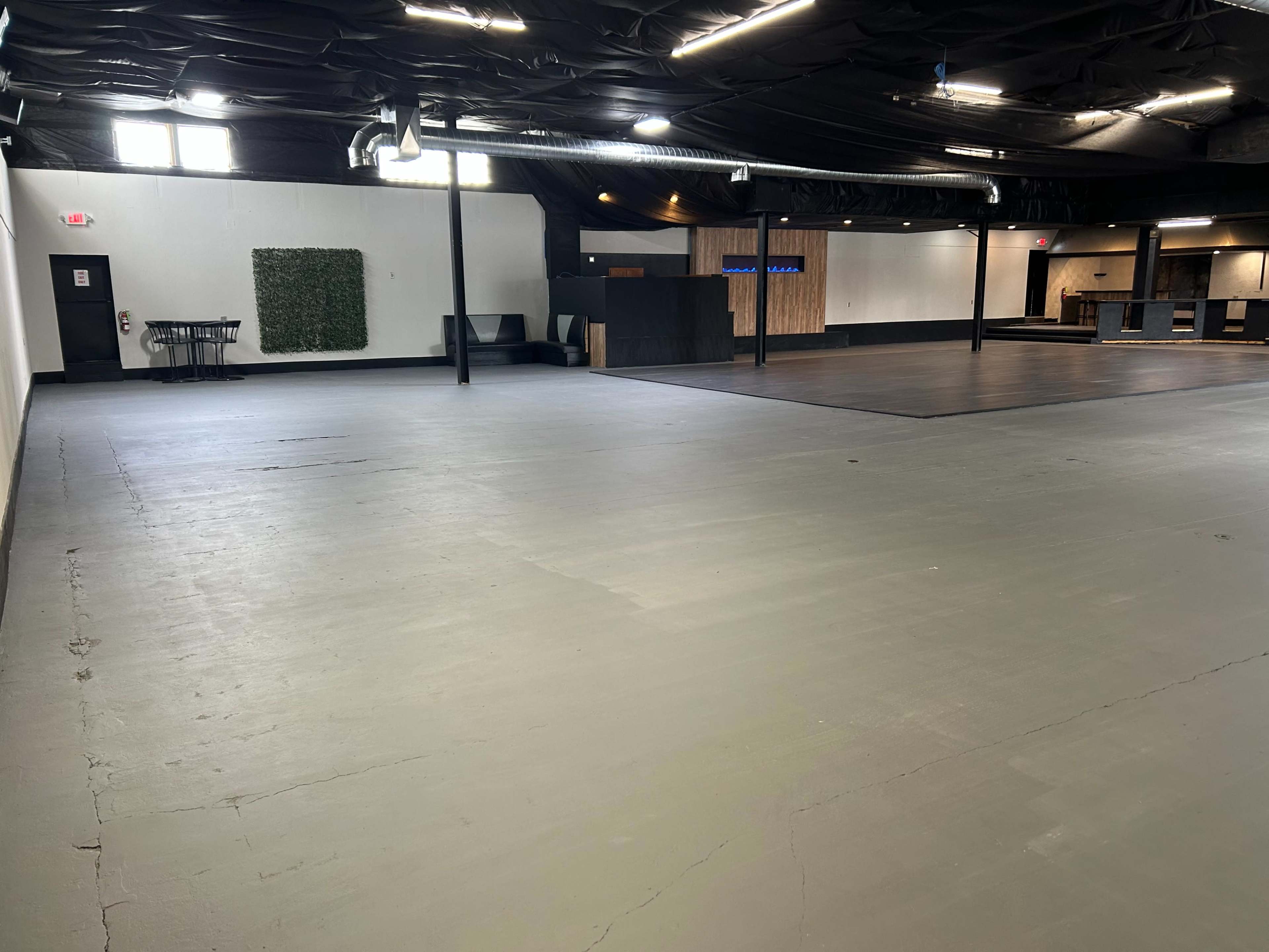 The image shows a spacious, empty room with a gray floor, black ceiling, and minimal furniture against the walls.