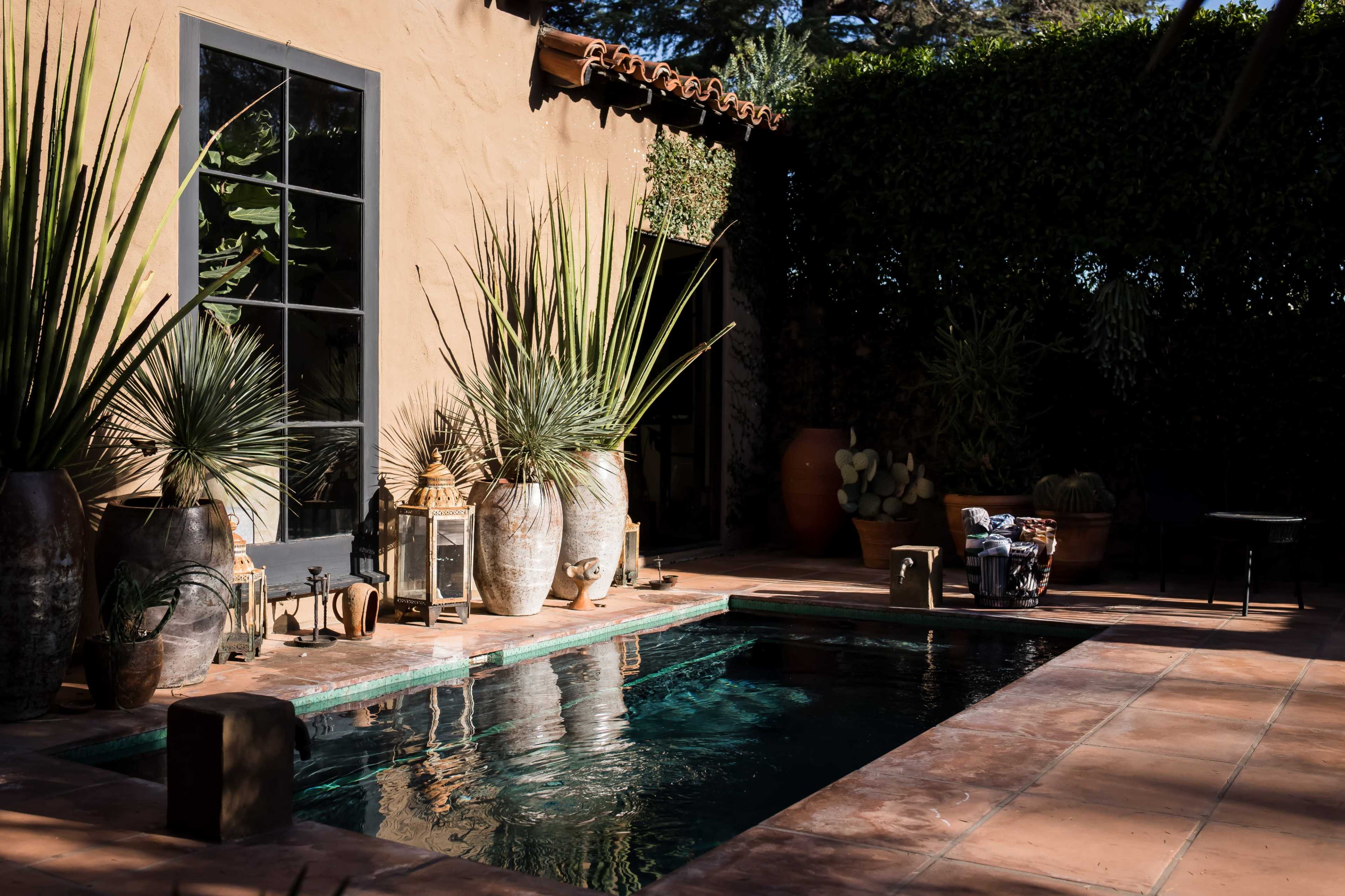 A tranquil outdoor space featuring a small pool surrounded by potted plants and decorative lanterns against a sunlit wall.