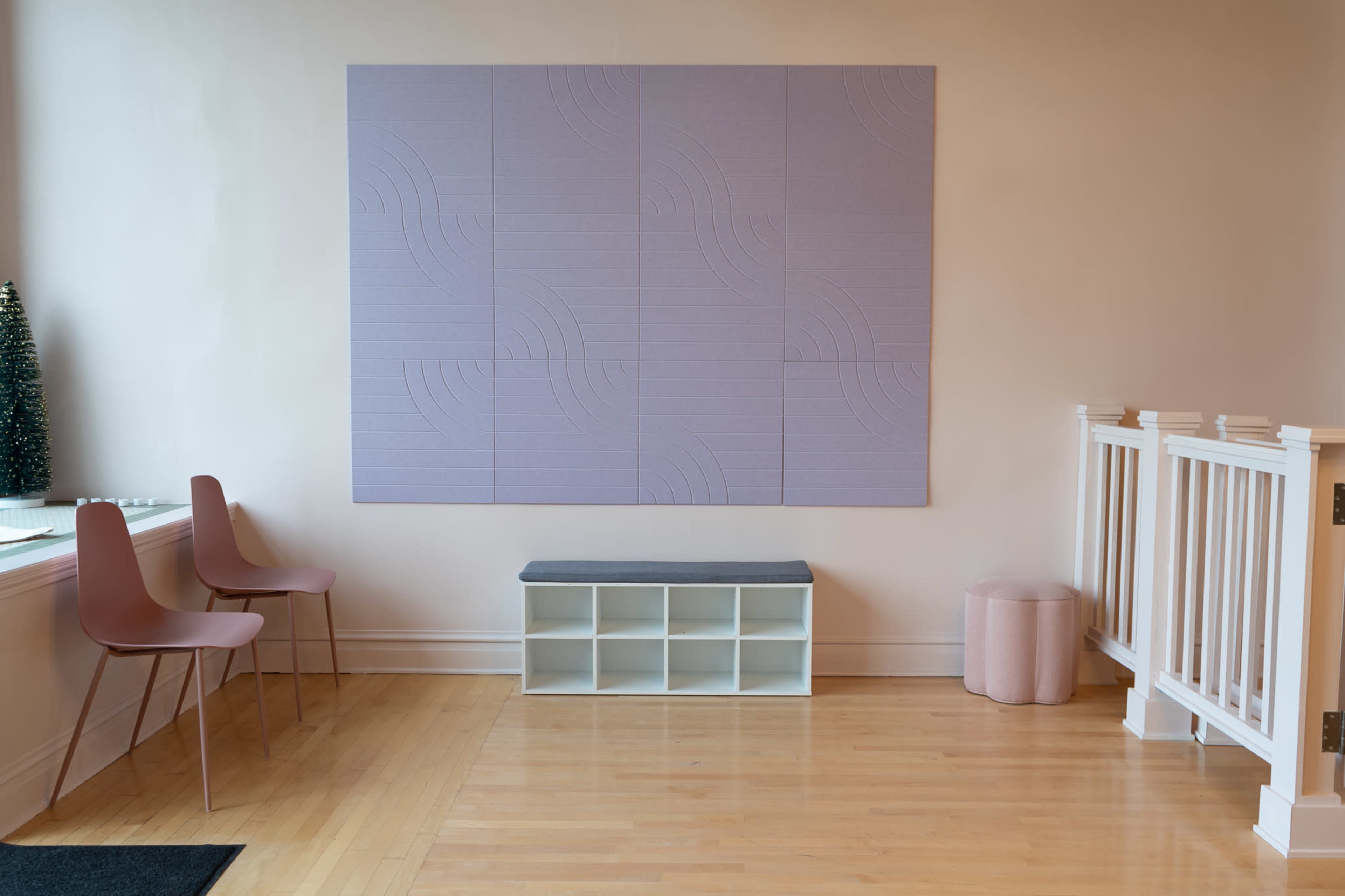 The image shows a minimalist room featuring a pale purple wall panel, two pink chairs, a white cubby bench, and a small cylindrical stool.
