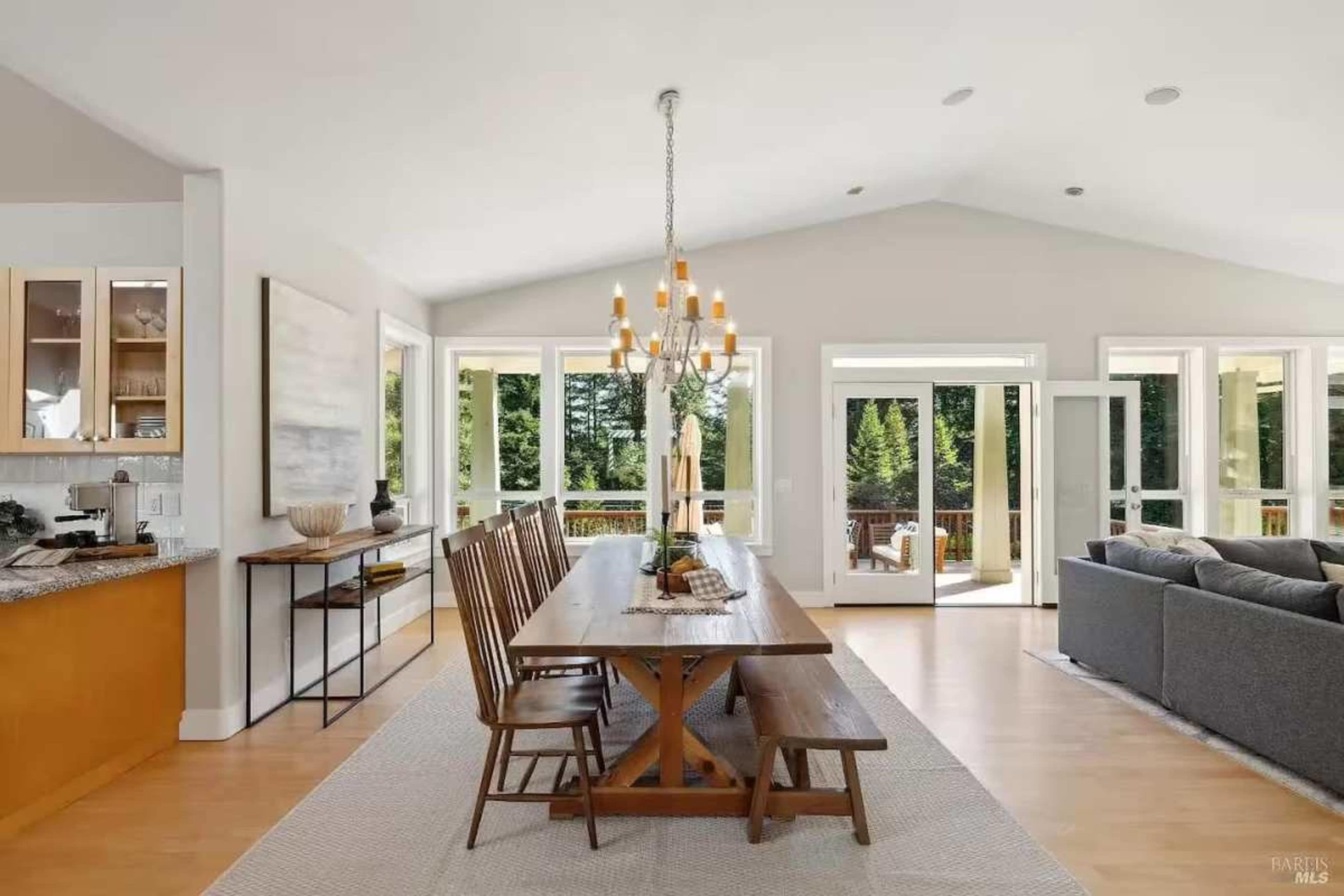 A spacious dining area features a long table with wooden chairs, surrounded by large windows that overlook a forested outdoor space.