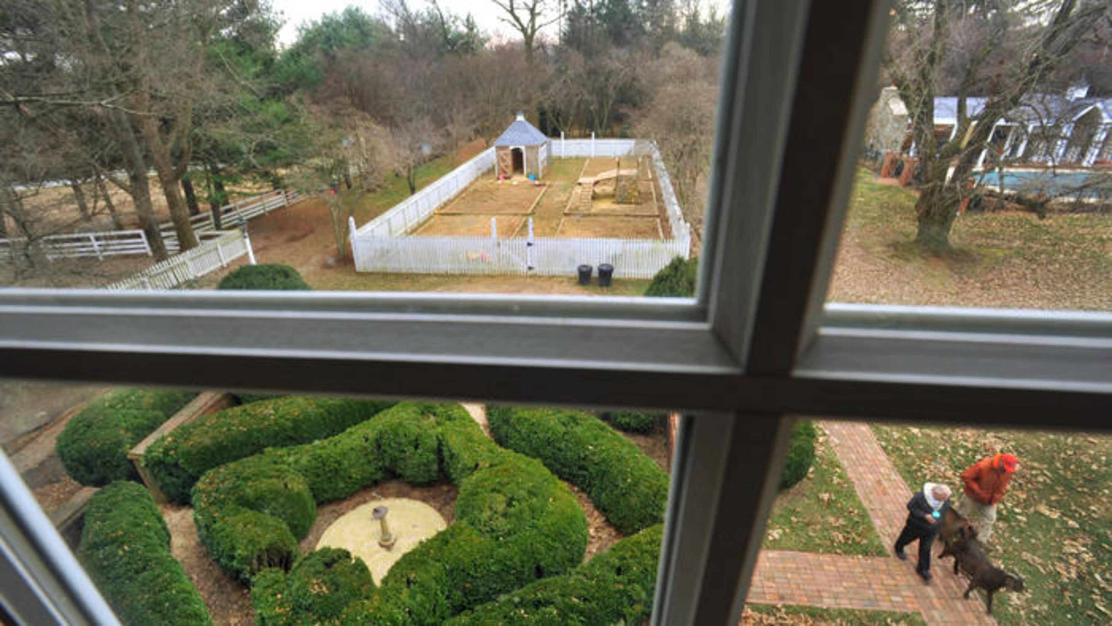 The view from a window shows a landscaped yard with a circular hedge, a fountain, and two people walking a dog near a fenced garden area.