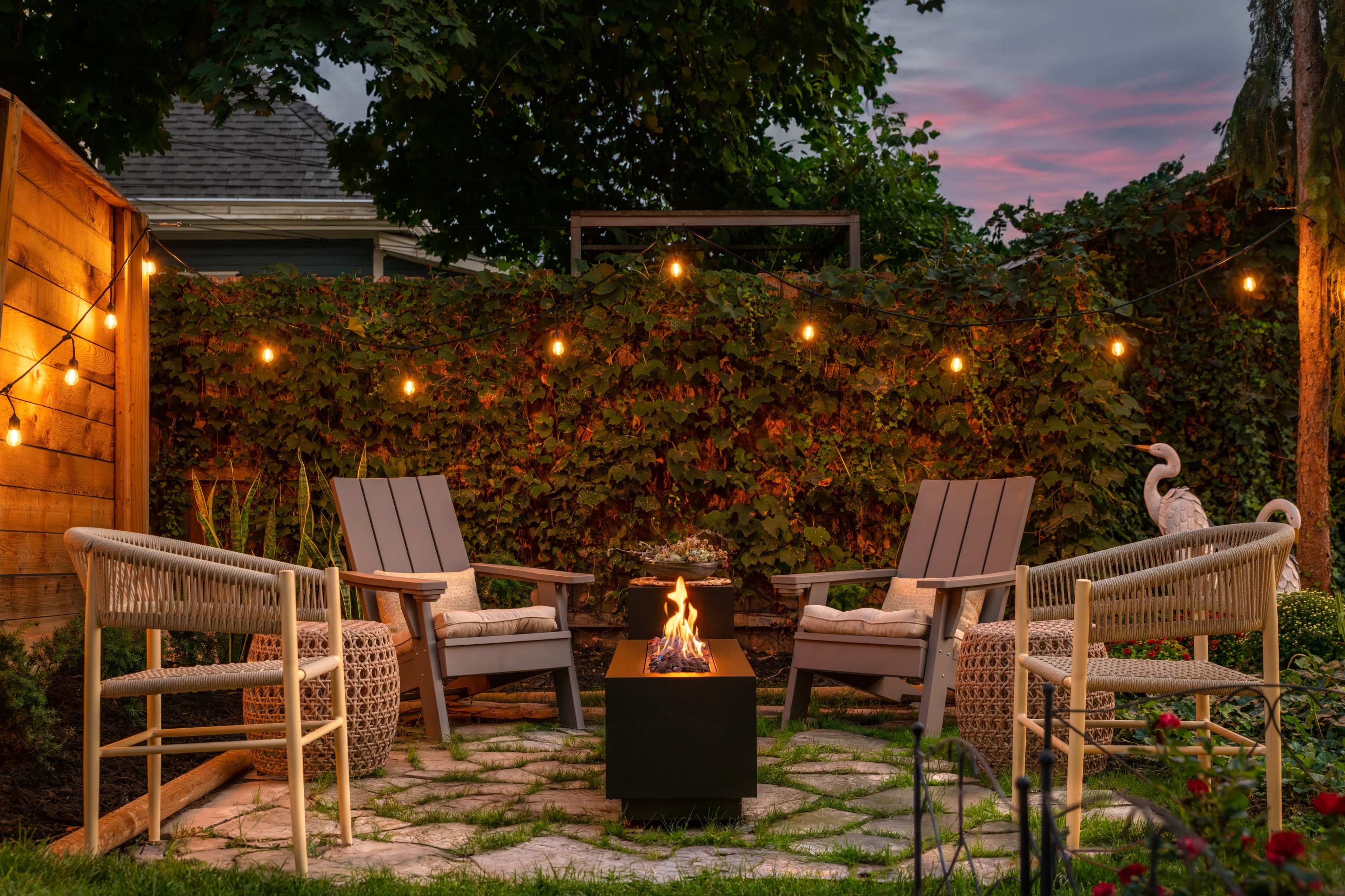 A cozy outdoor seating area features two chairs facing a fire pit surrounded by lush greenery and string lights.