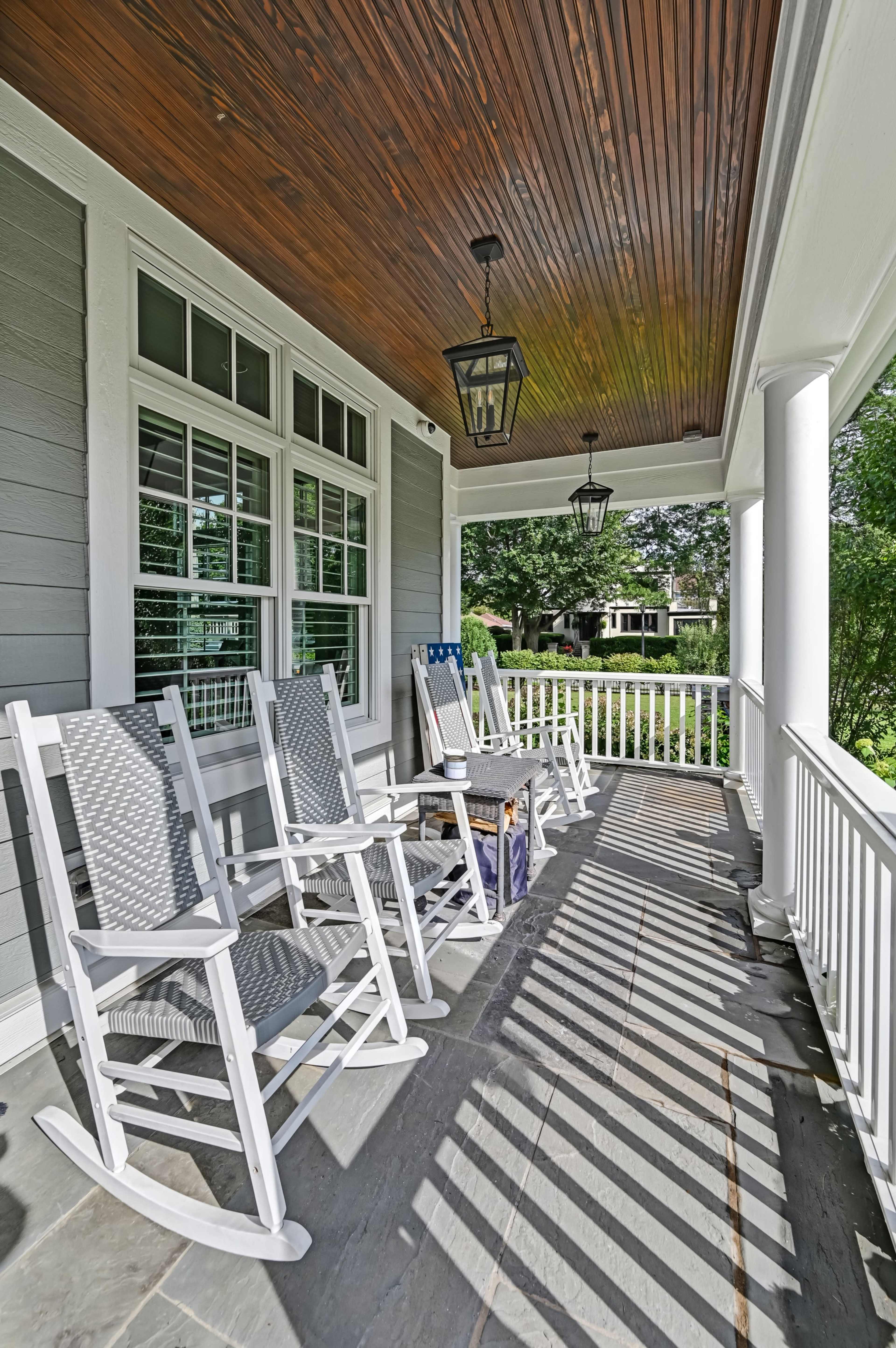 The image depicts a porch with white rocking chairs, a small table, and overhead lanterns, surrounded by greenery and sunlight casting shadows on the stone floor.