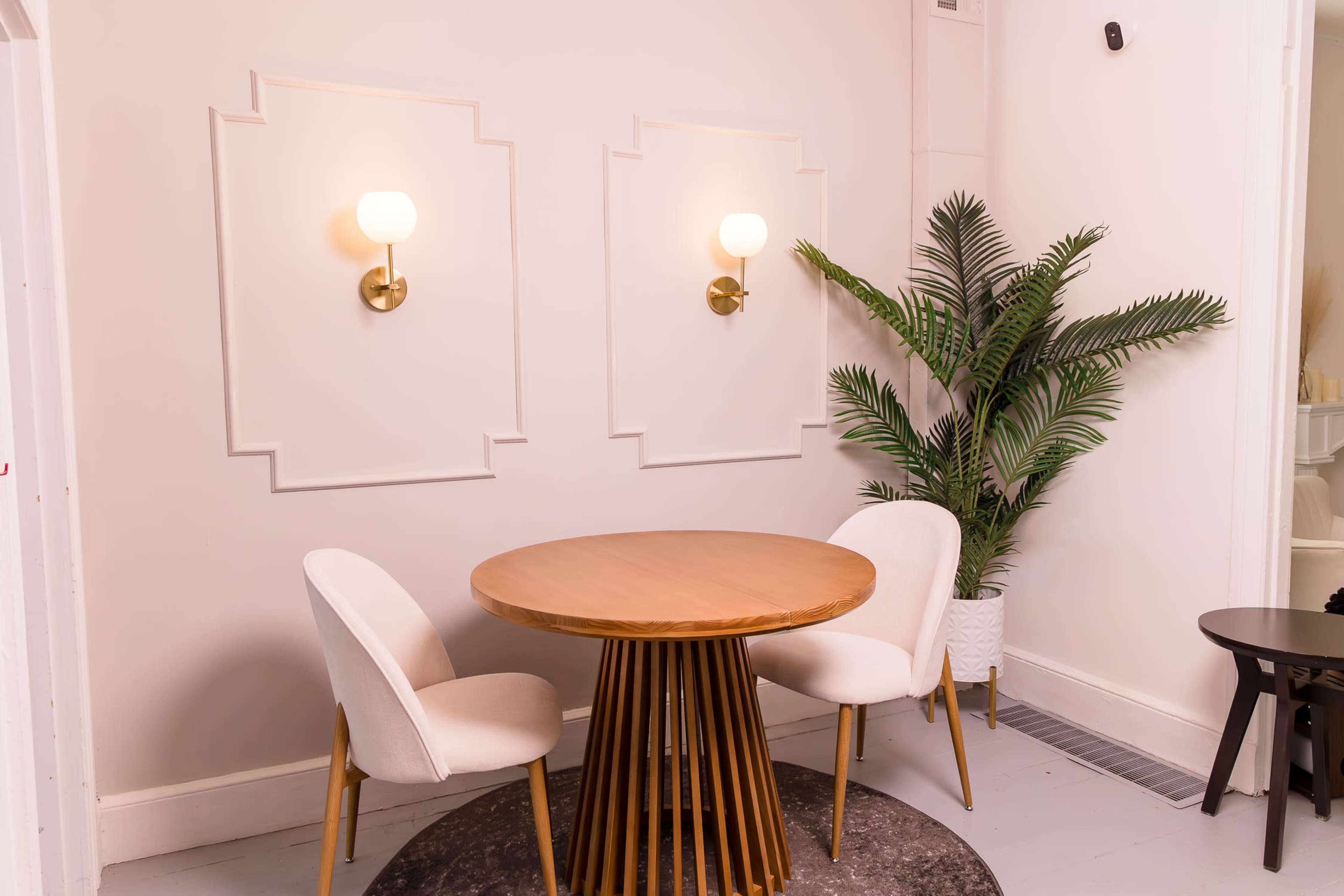 The image features a small circular wooden table with two upholstered chairs beside it, set against a pale wall with decorative sconce lighting and a potted plant.