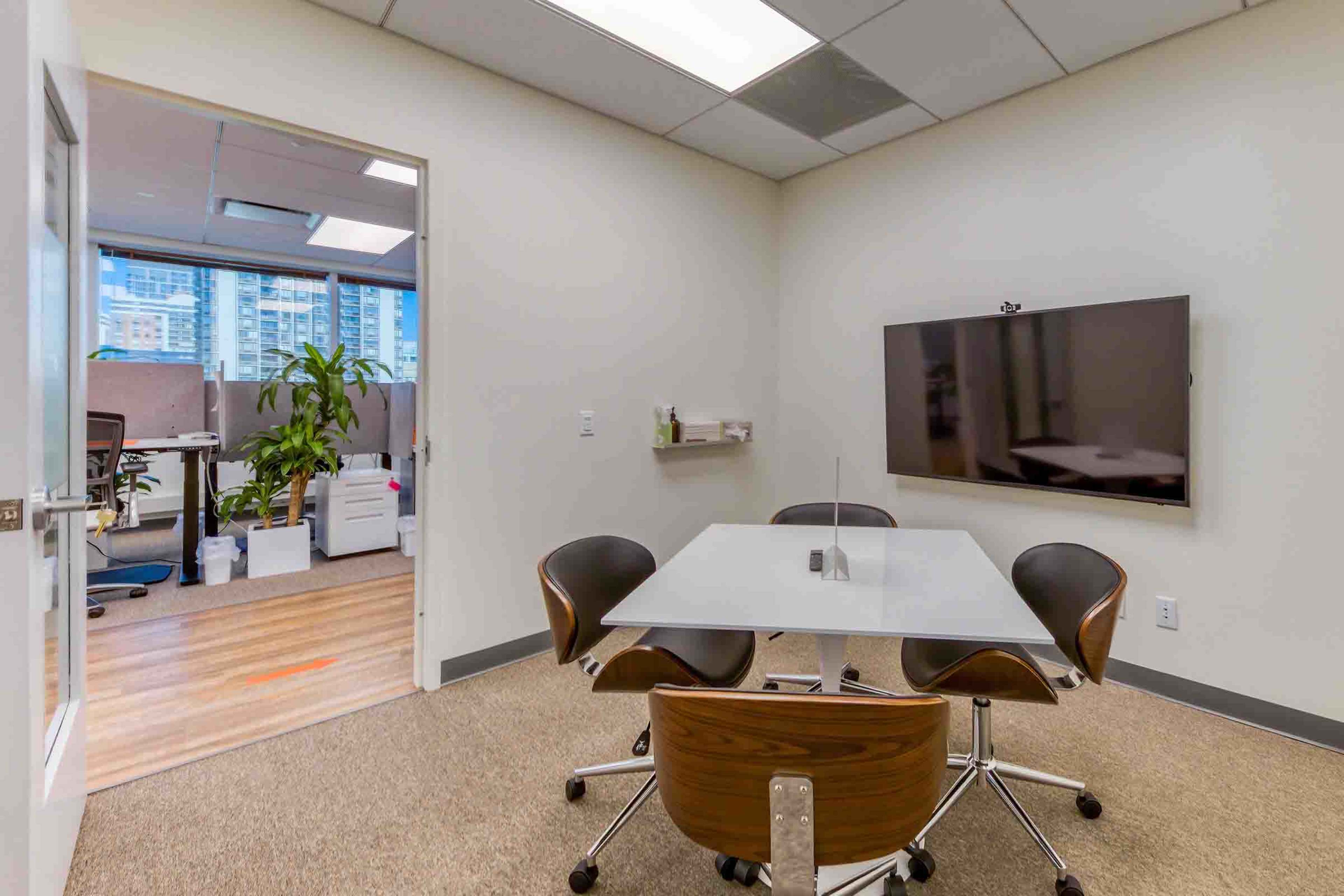 A small meeting room features a white table surrounded by four brown chairs, with a television mounted on the wall and an open office area visible through the doorway.