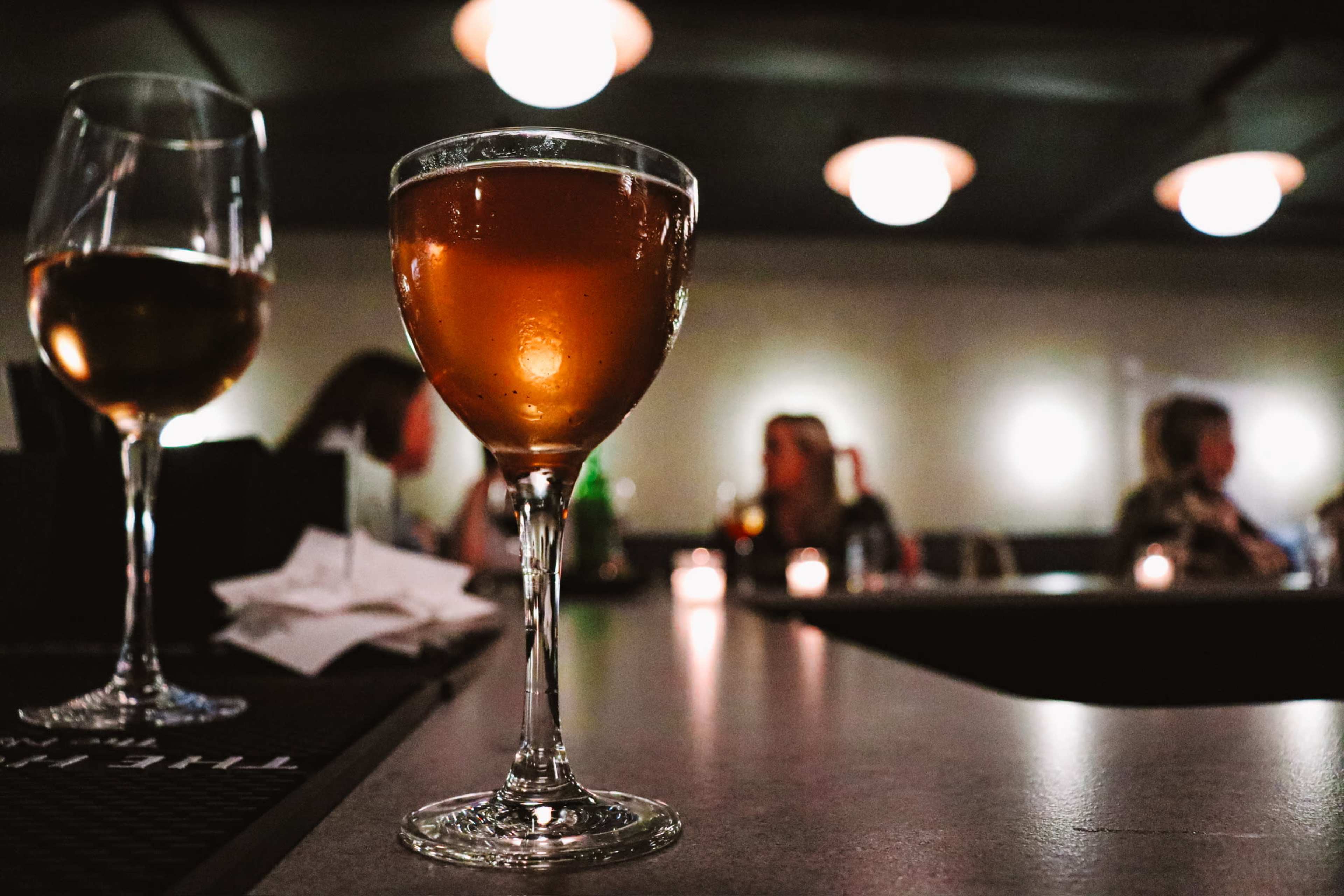 A clear cocktail glass filled with amber liquid sits on a bar counter in the foreground, with blurred patrons socializing in the background.