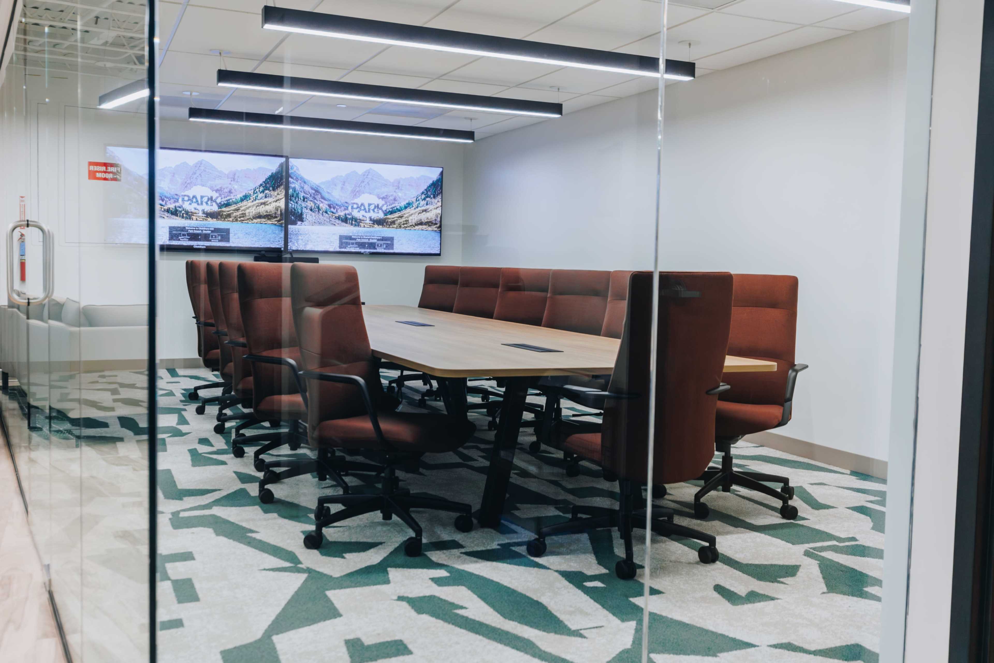 A modern conference room features a large rectangular table surrounded by ergonomic chairs, with two large screens displaying mountain scenery on the walls.