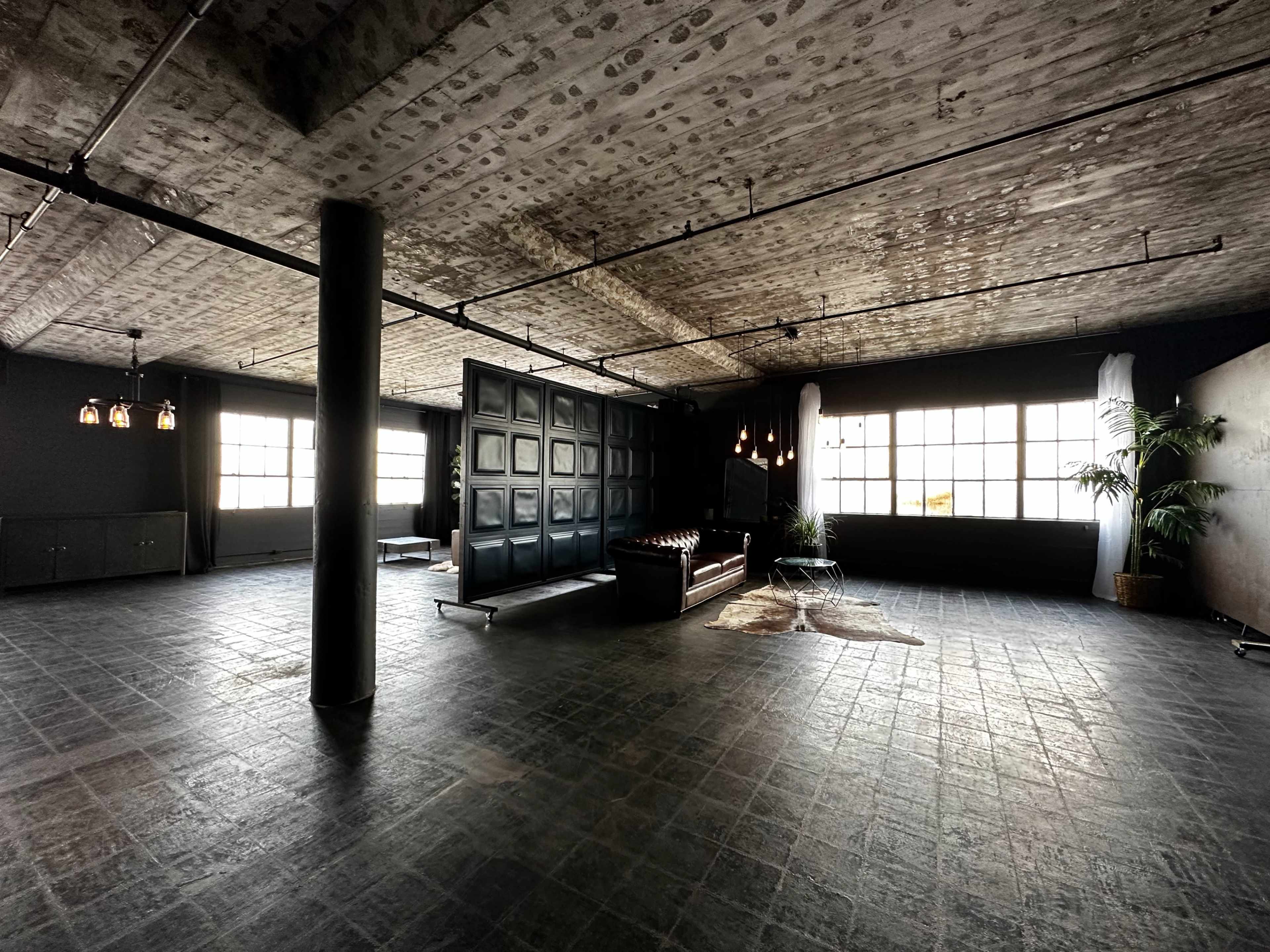 The image shows a spacious, industrial-style room with large windows, a leather sofa, and minimal furnishings.