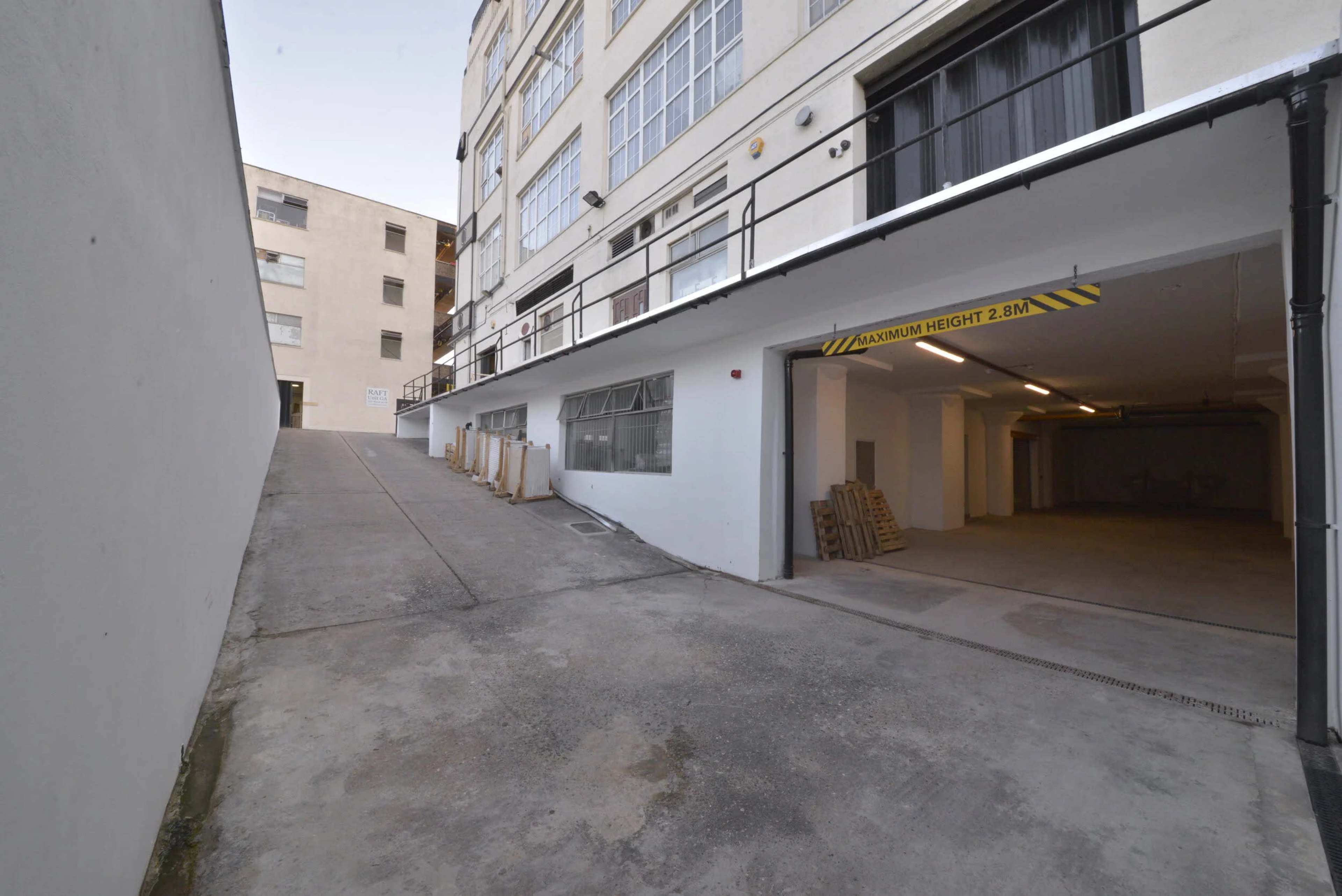 The image shows a sloped driveway leading to a parking garage entrance beneath a building, with wooden pallets stacked nearby.