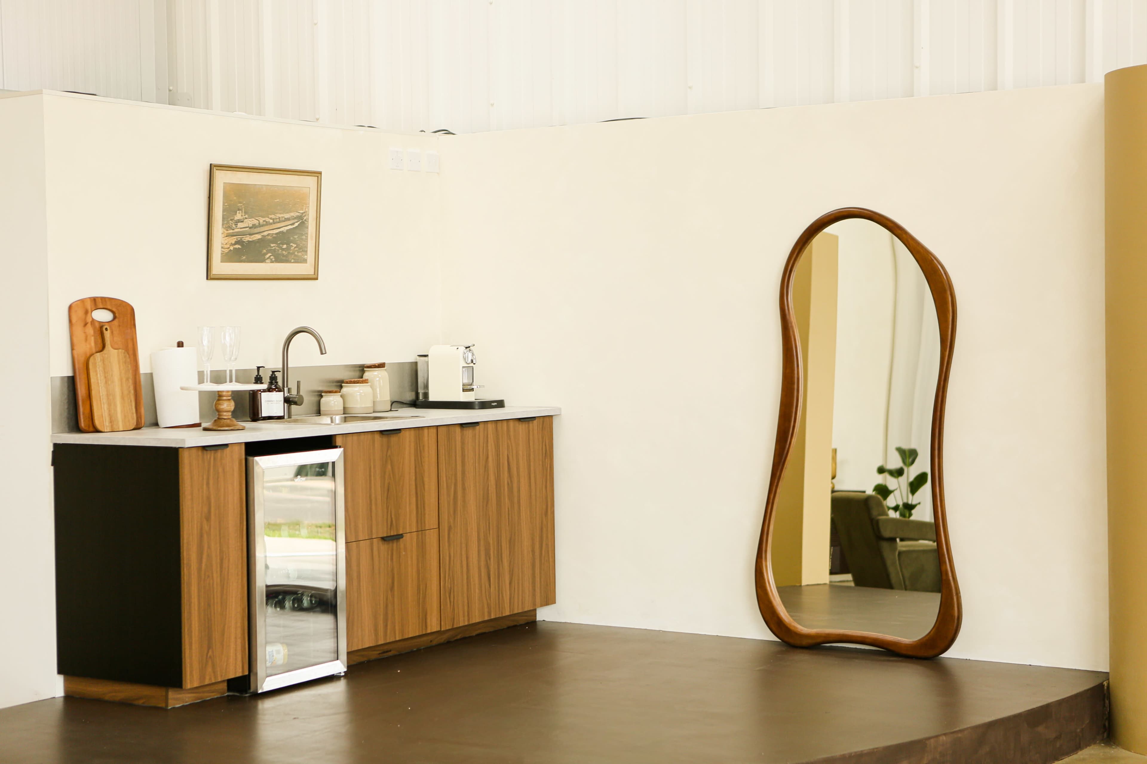 The image shows a modern kitchen area with a wooden cabinet, sink, and a large, curvy mirror leaning against the wall.