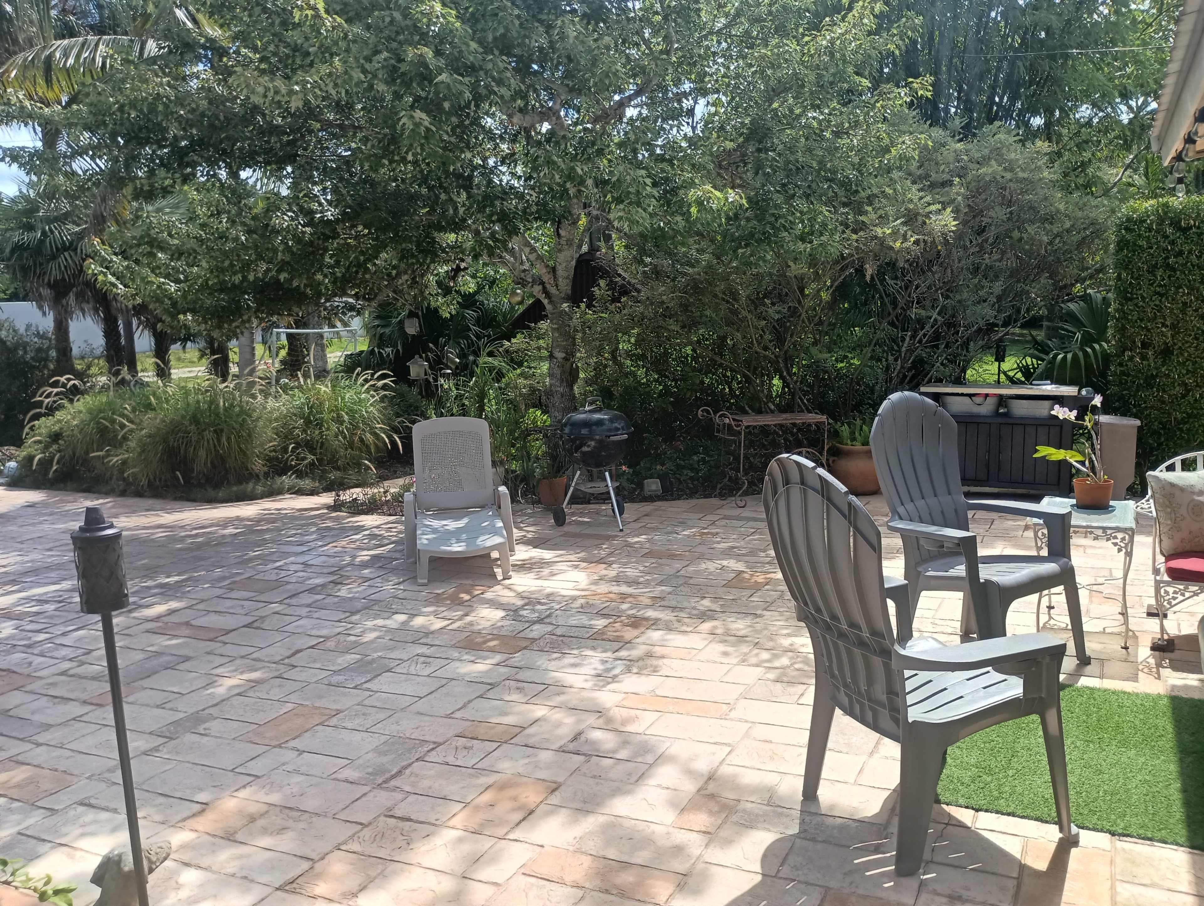 The image shows a stone patio with several outdoor chairs, a small grill, and surrounded by greenery and trees.