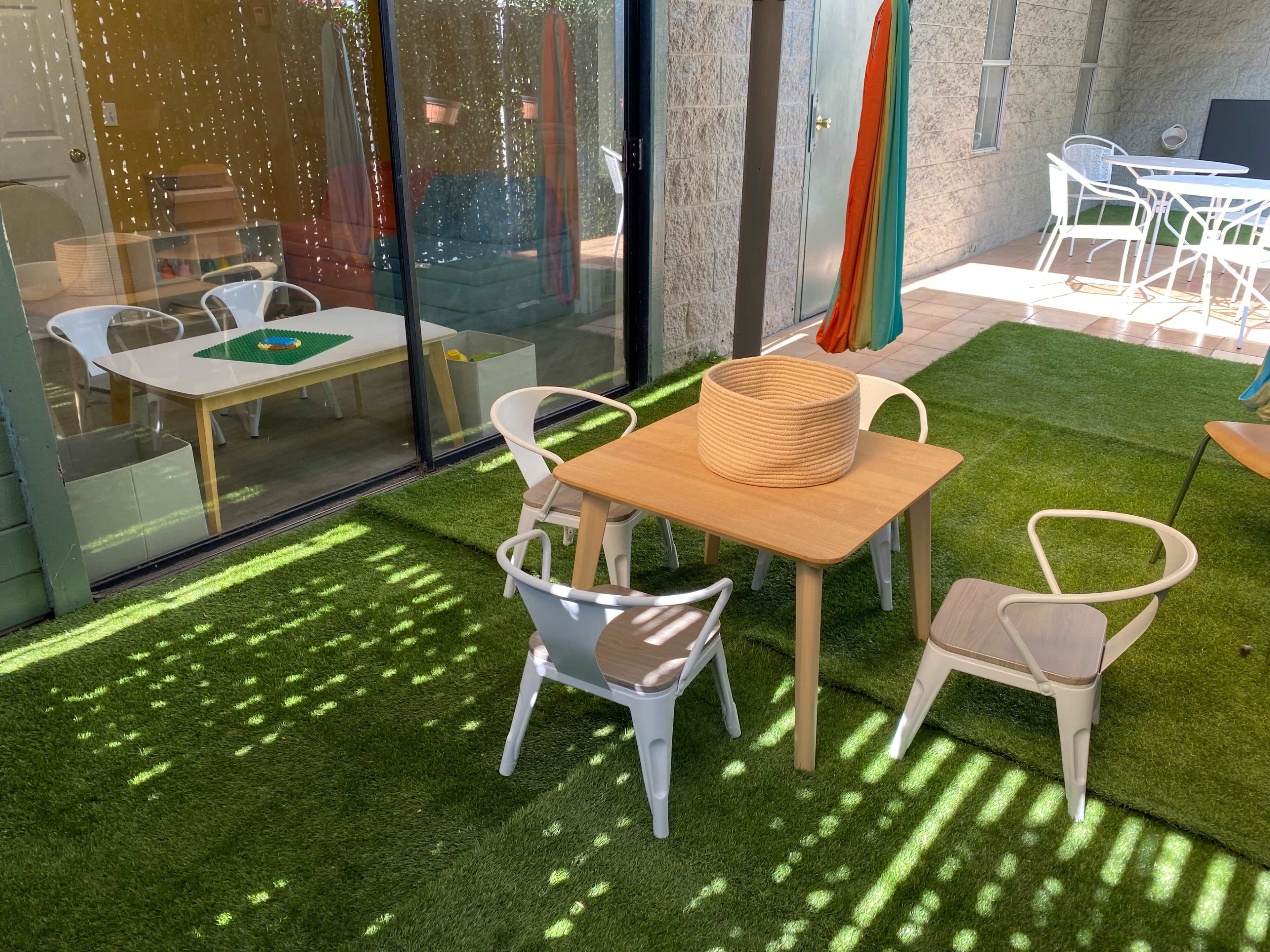The image shows a small outdoor area with a wooden table surrounded by five chairs, set on artificial grass, with colorful curtains and reflections in the windows.