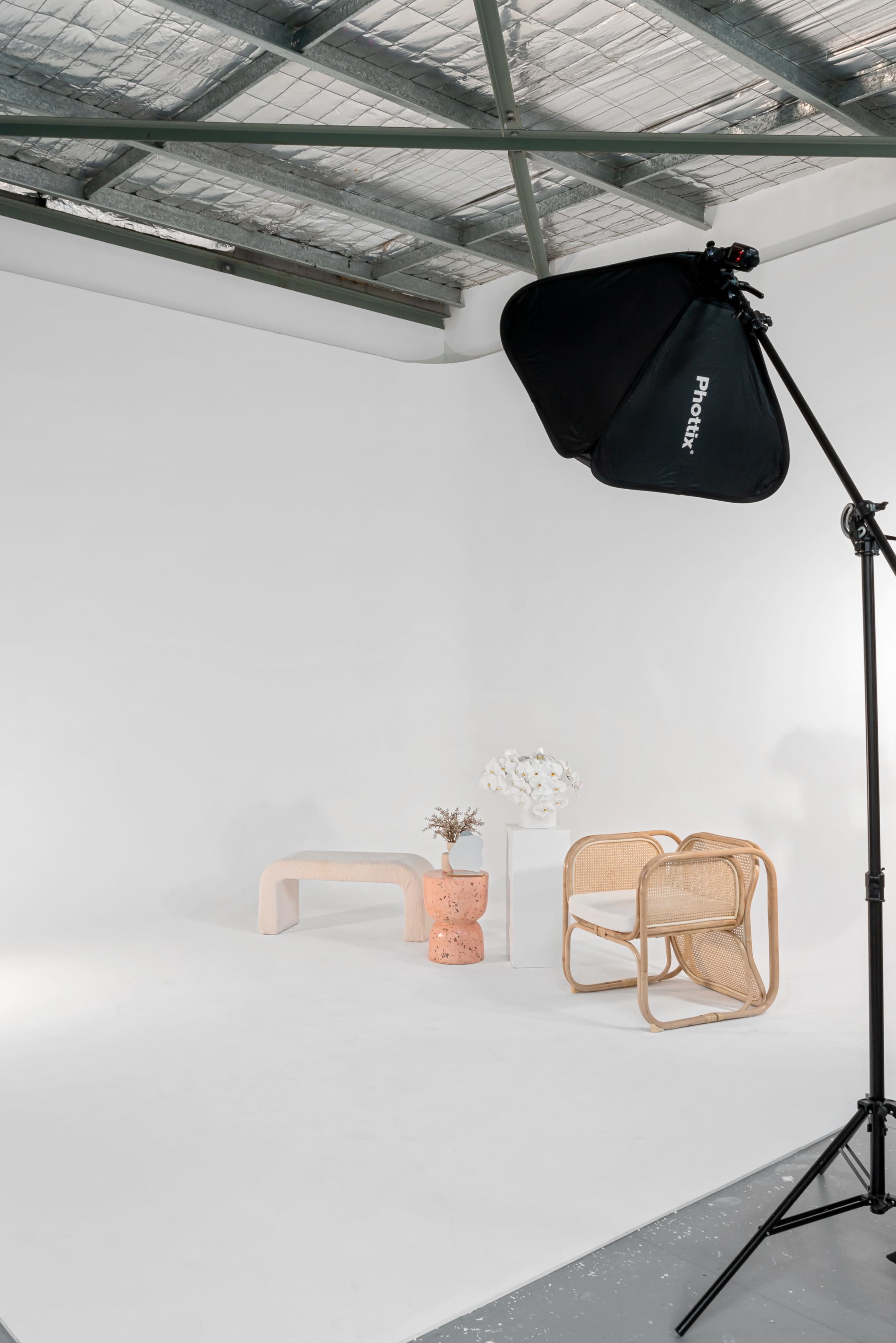 A photography studio features minimalist furniture, including a curved wooden chair and a small table, with a softbox light set up on the left.