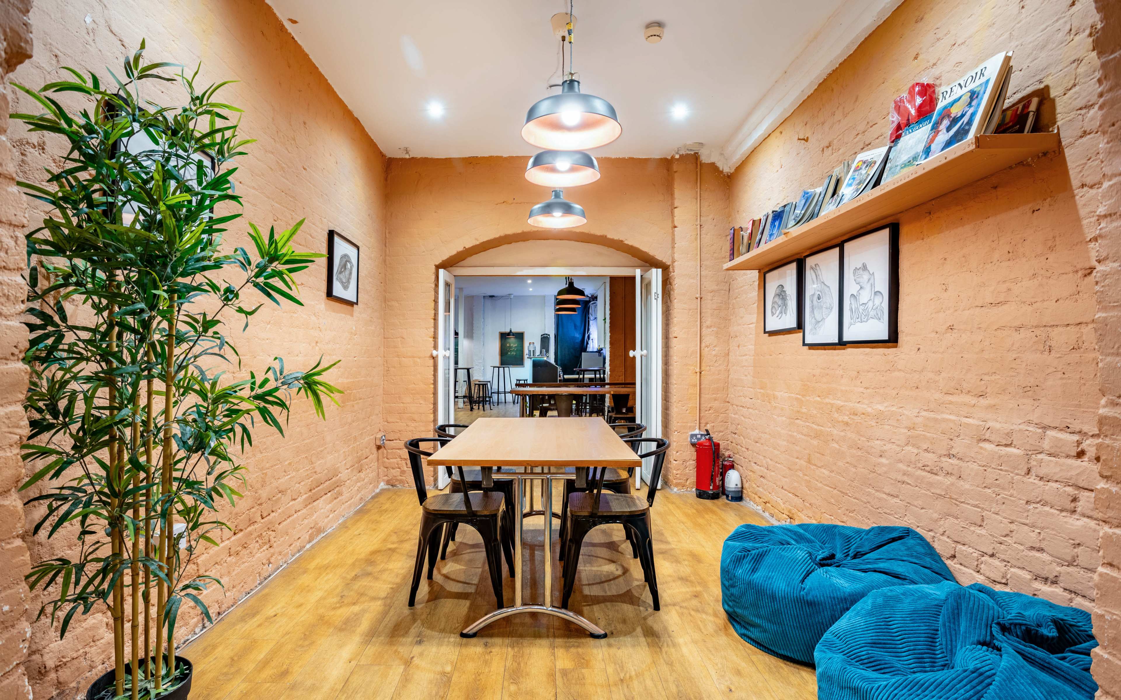 A workspace with a wooden table and four black chairs, flanked by a plant and shelves holding books and framed art on the walls.