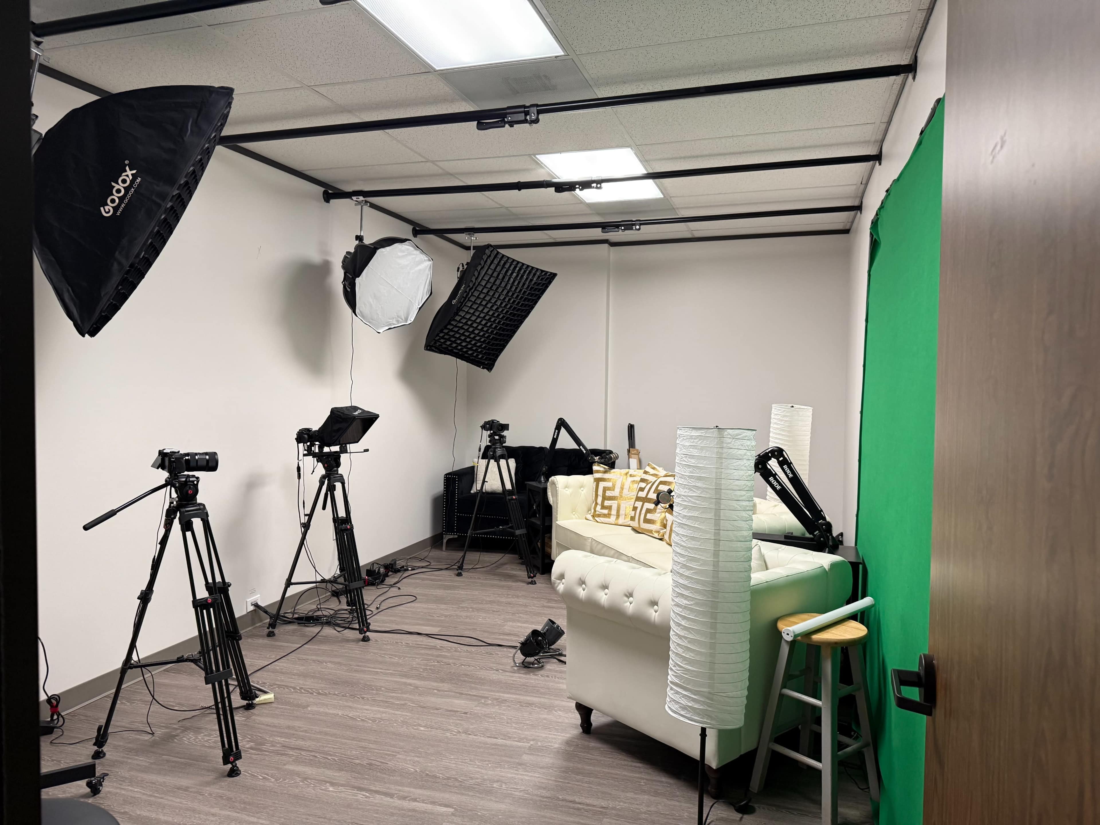 The image shows a studio setup with multiple cameras, lighting equipment, and a green screen arranged in a small room.