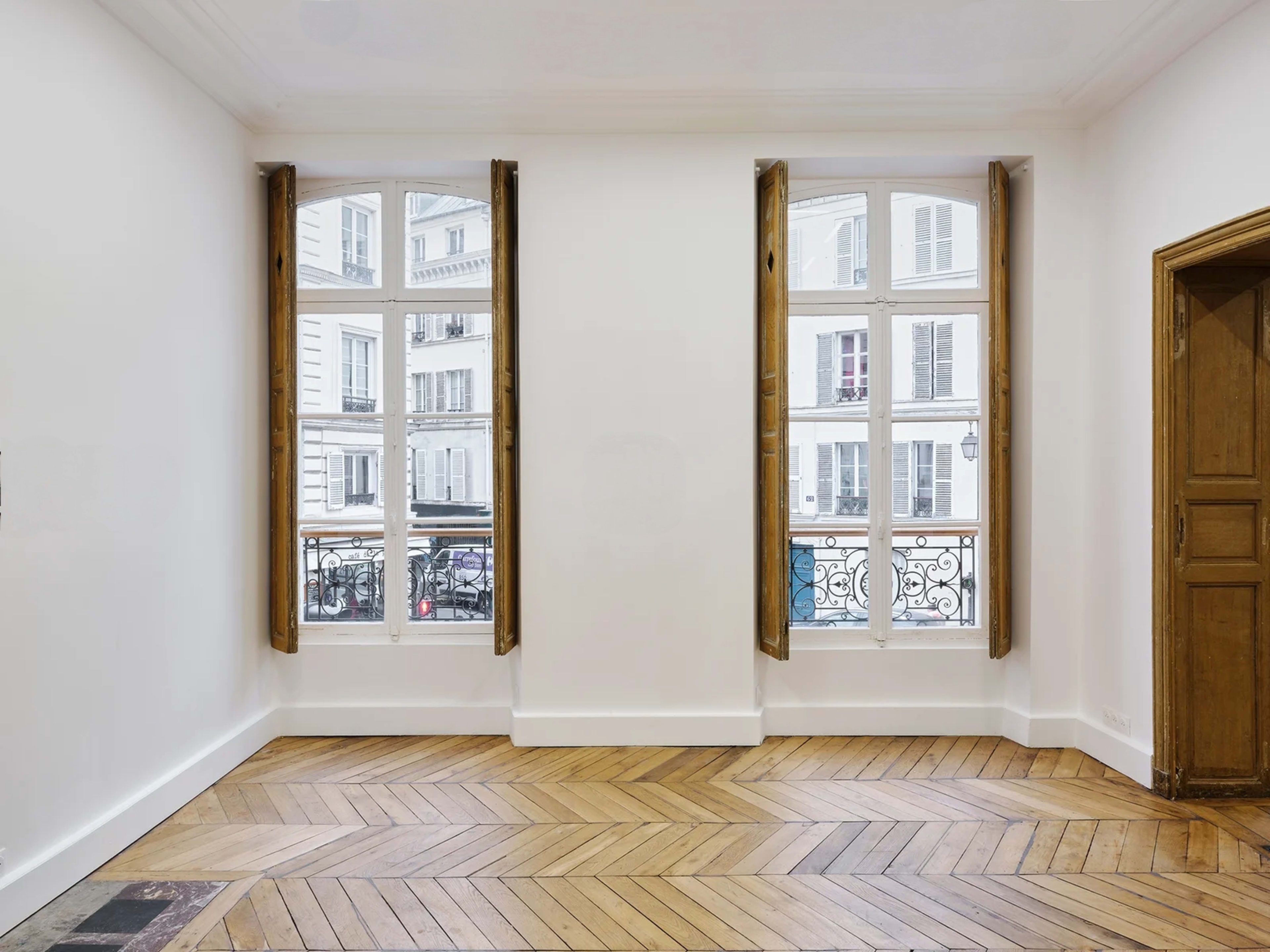 The room features two tall windows with wooden shutters, showcasing a view of a street through ornate iron railings, while the herringbone wood floor adds texture to the space.