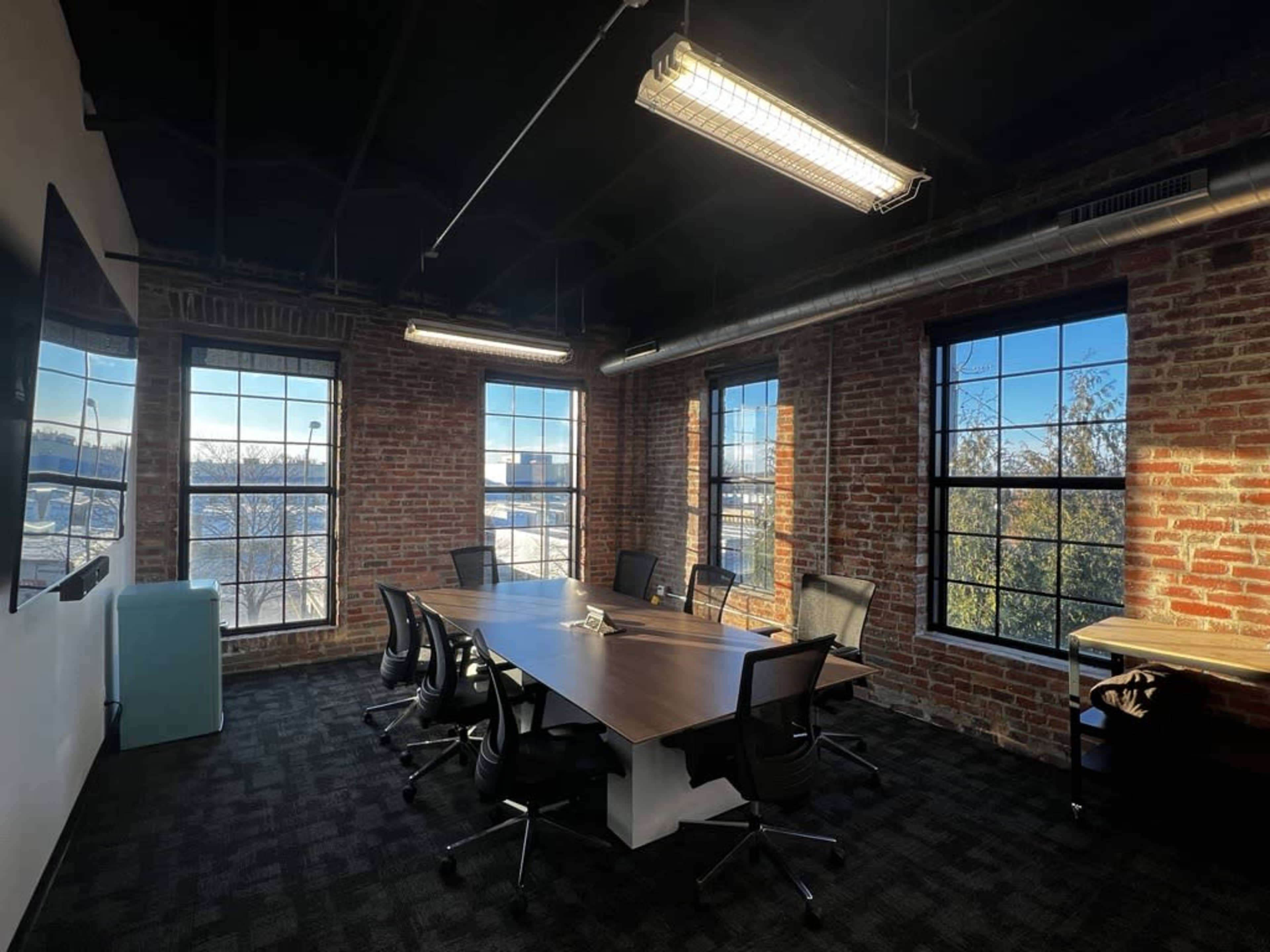 A conference room with a large wooden table surrounded by black chairs, featuring exposed brick walls and large windows that let in natural light.