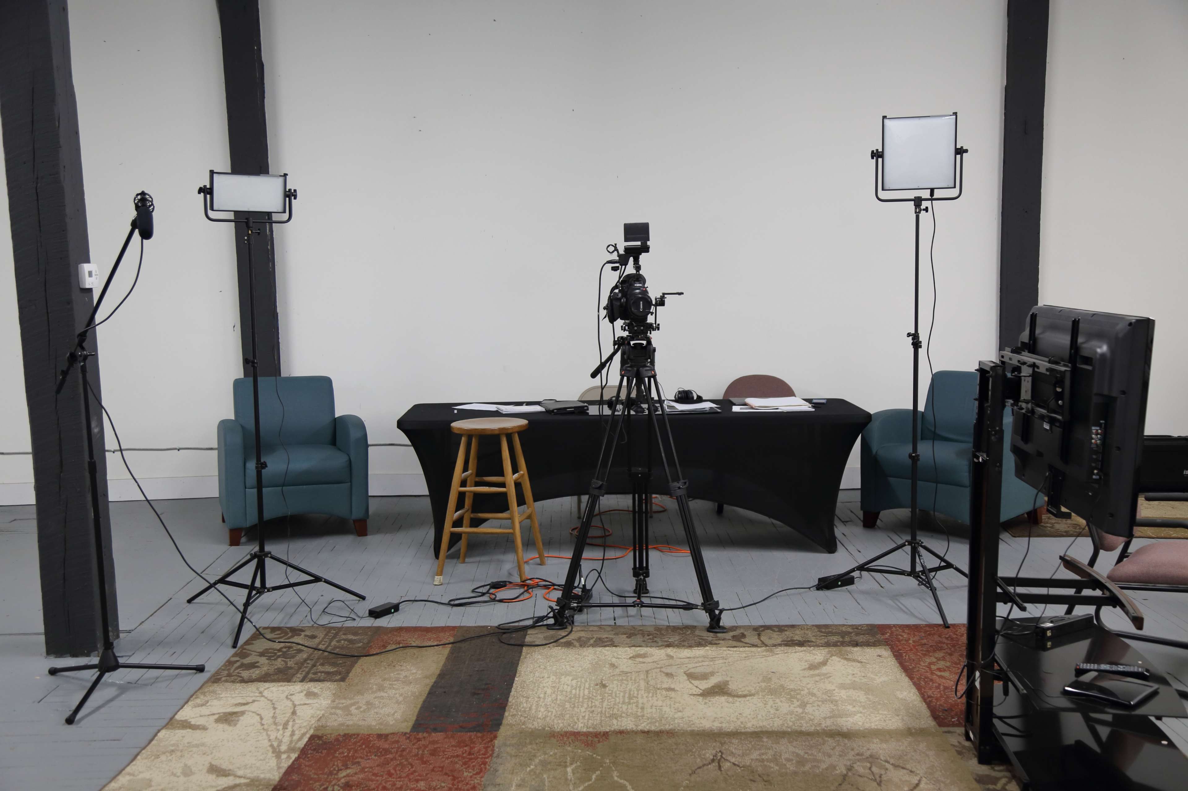 A video recording setup features a table, two chairs, and multiple cameras and lights arranged in a spacious room with a light-colored floor and a patterned rug.