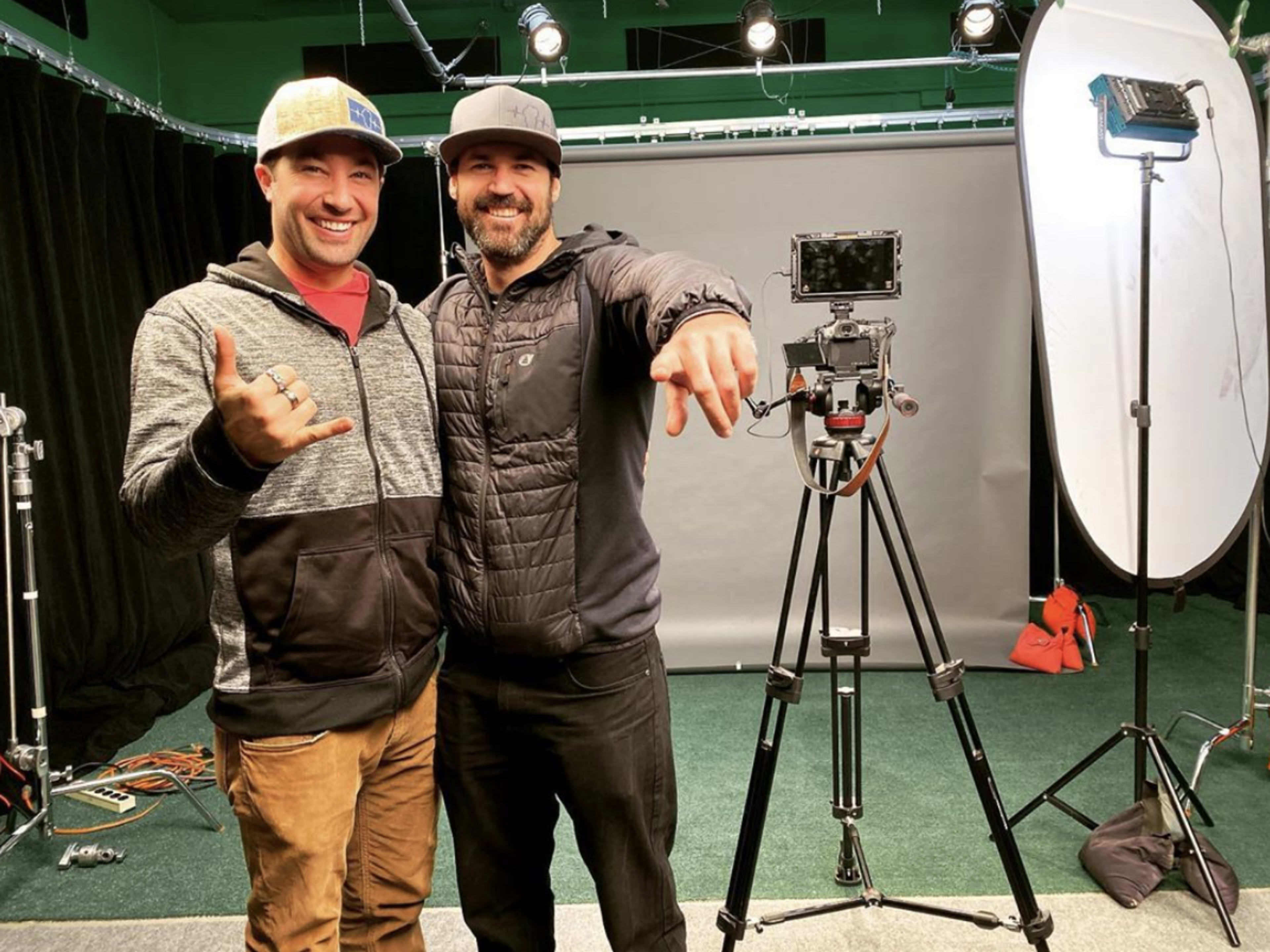 Two men pose together in a studio setting, smiling for the camera with a video camera and lighting equipment in the background.