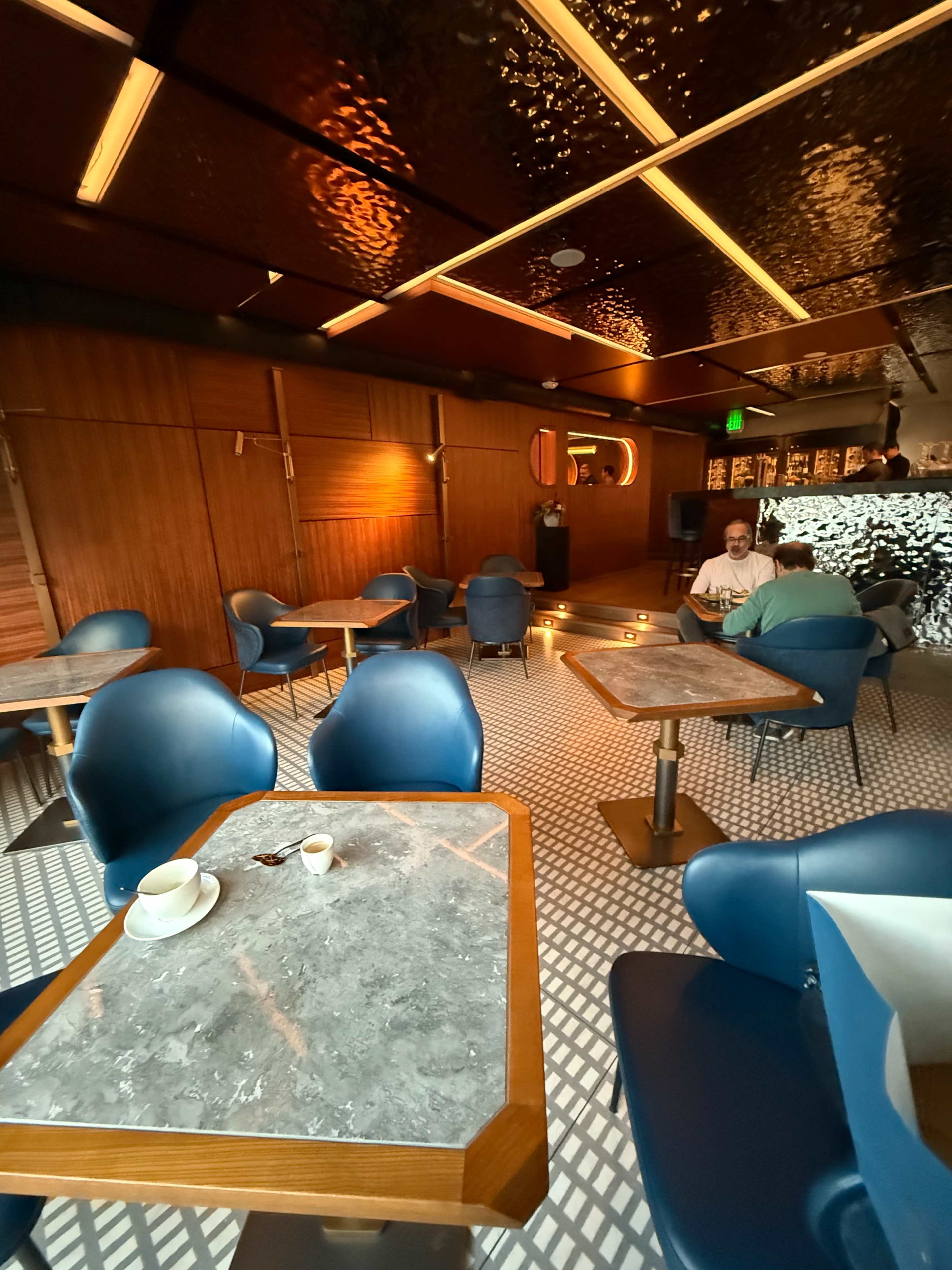 The image shows a modern café interior with wooden walls, sleek blue chairs, and marble-topped tables, illuminated by soft ambient lighting.