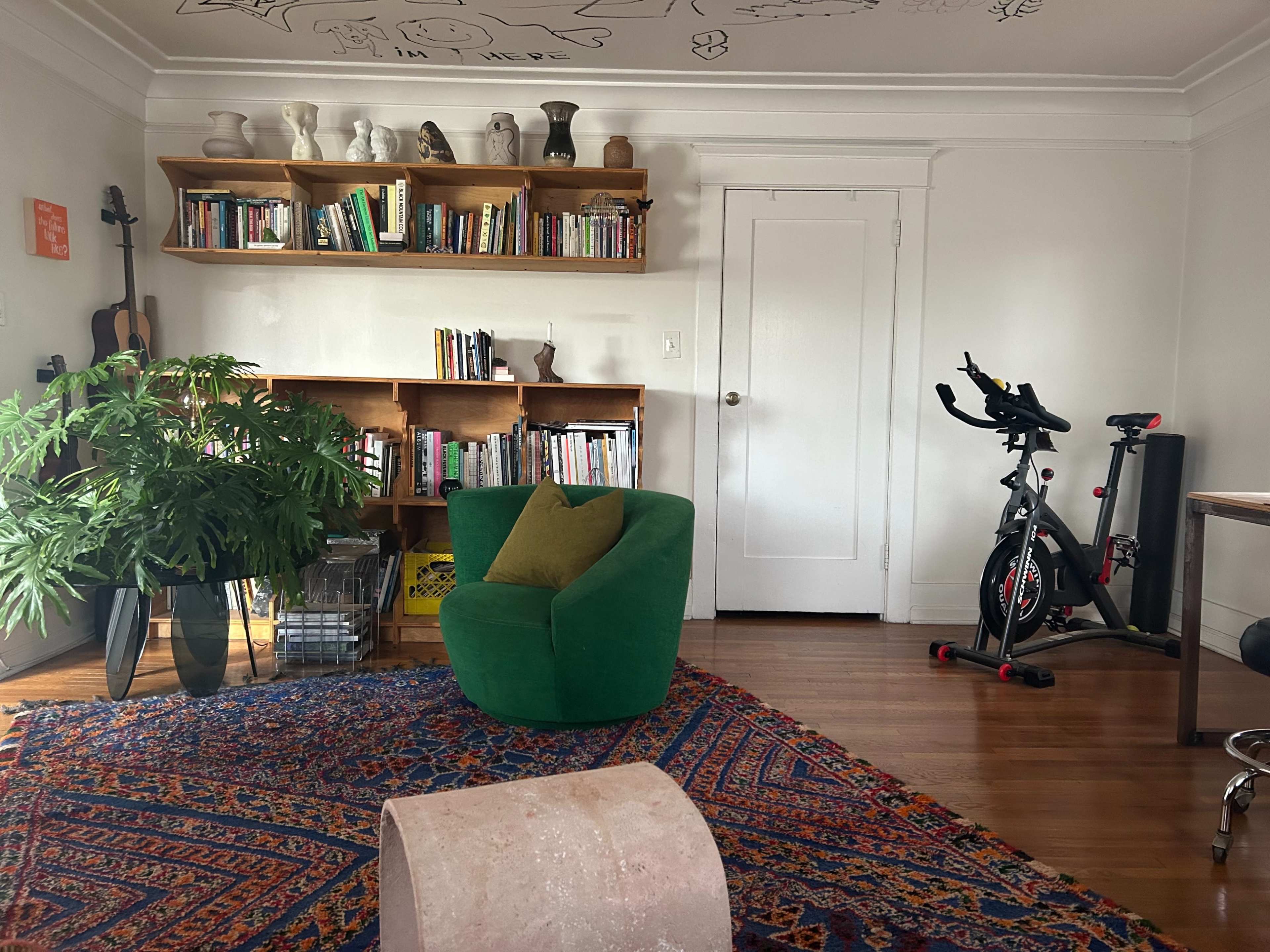 The scene features a cozy living room with a green chair, bookshelves filled with books, a plant, a wall-mounted bike, and a patterned rug.