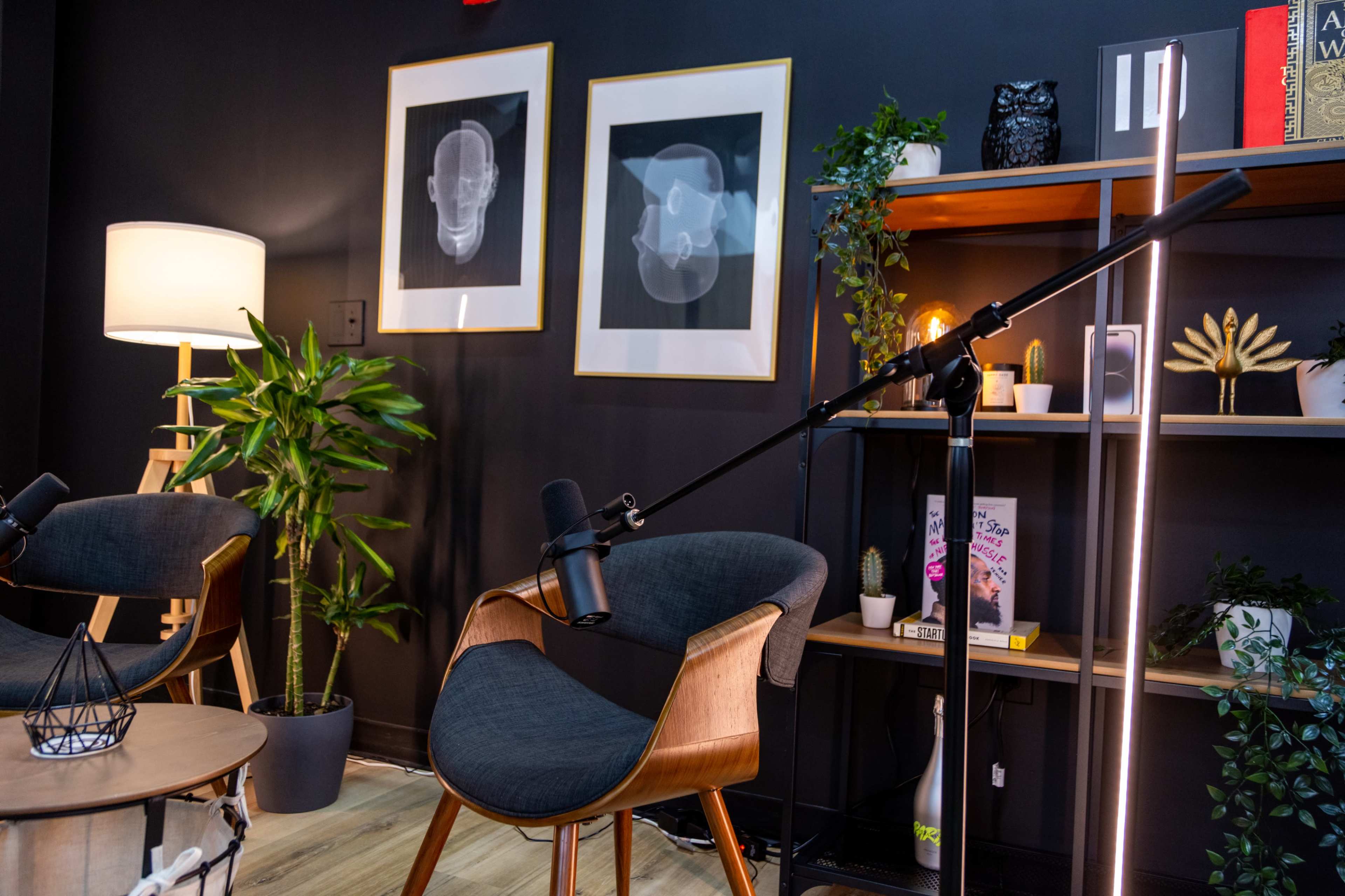 A cozy studio setup features two microphones positioned near stylish chairs, surrounded by plants and framed artwork on a dark wall.
