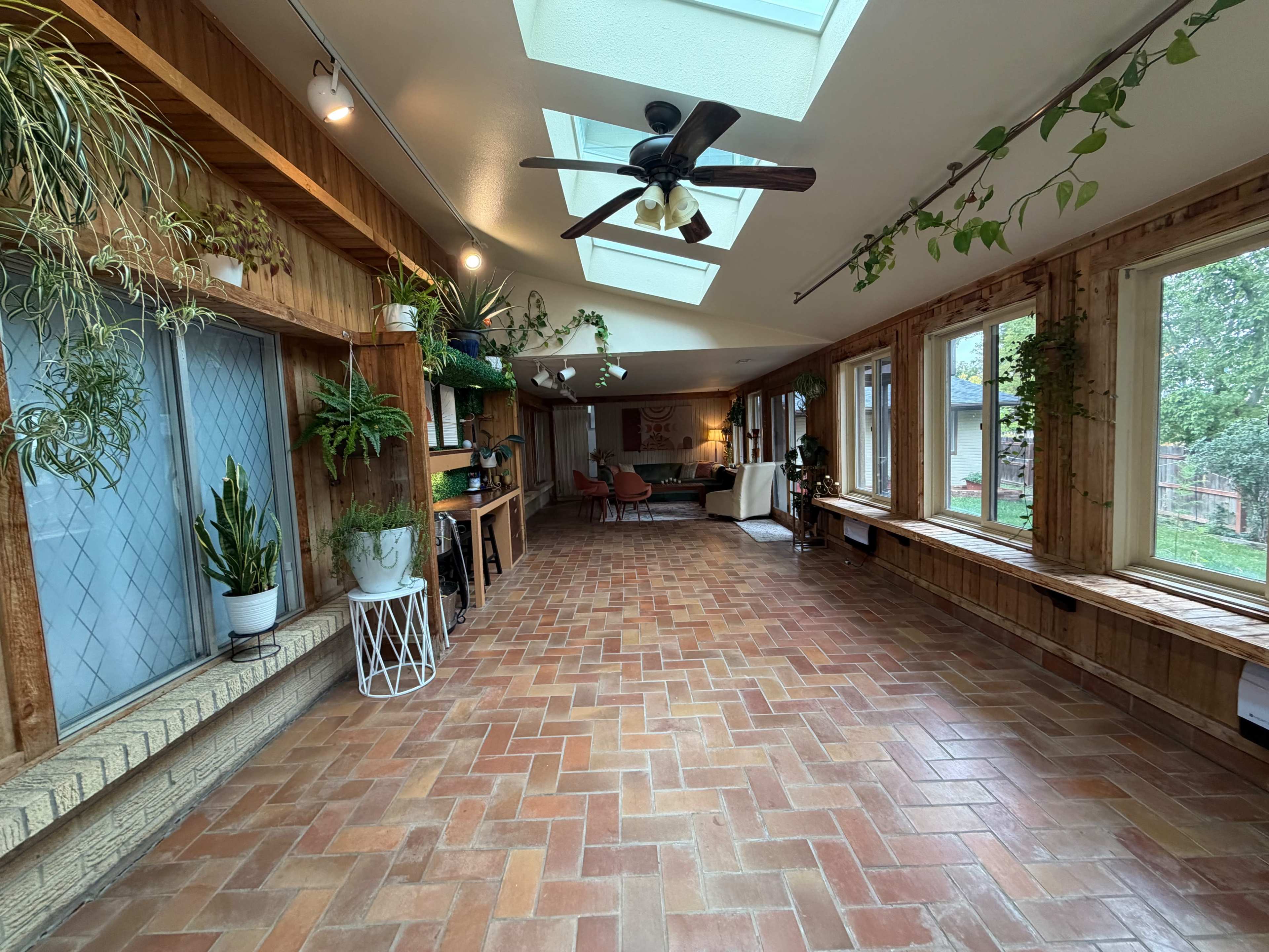 The image shows a spacious sunroom featuring brick flooring, large windows, and greenery along the walls.